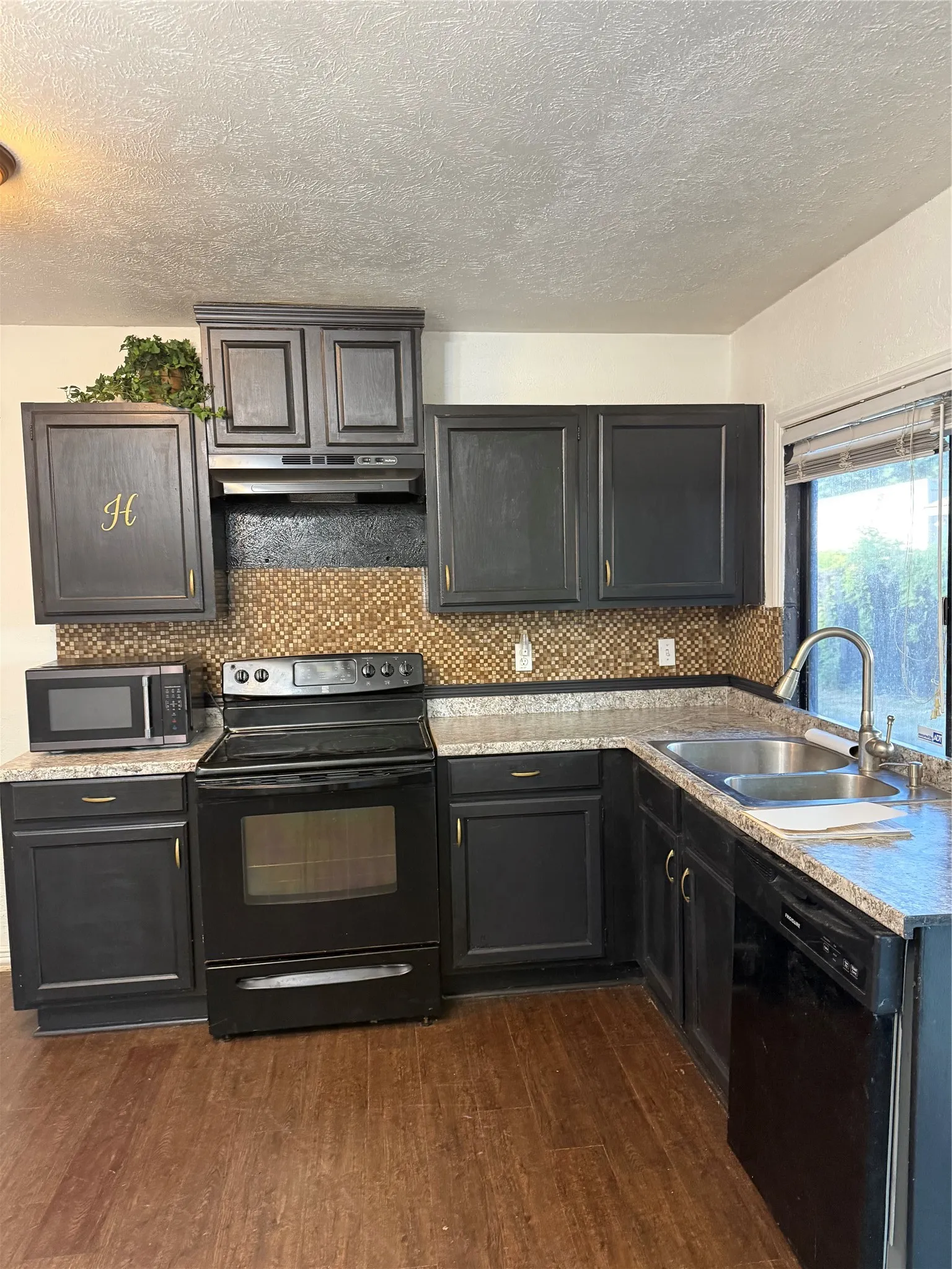 Kitchen featuring dark wood-finish cabinetry, mosaic tile backsplash, wood-finish flooring, stainless steel double basin sink, and black appliances