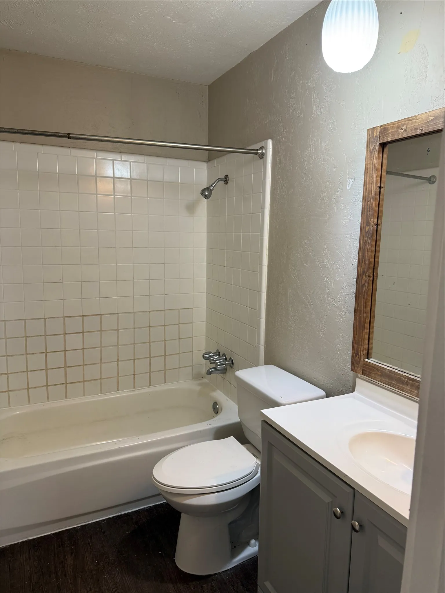 Bathroom featuring a tile-surround shower-tub combination with a showerhead, a toilet, a vanity with a white countertop and integrated sink, and wood-finish flooring