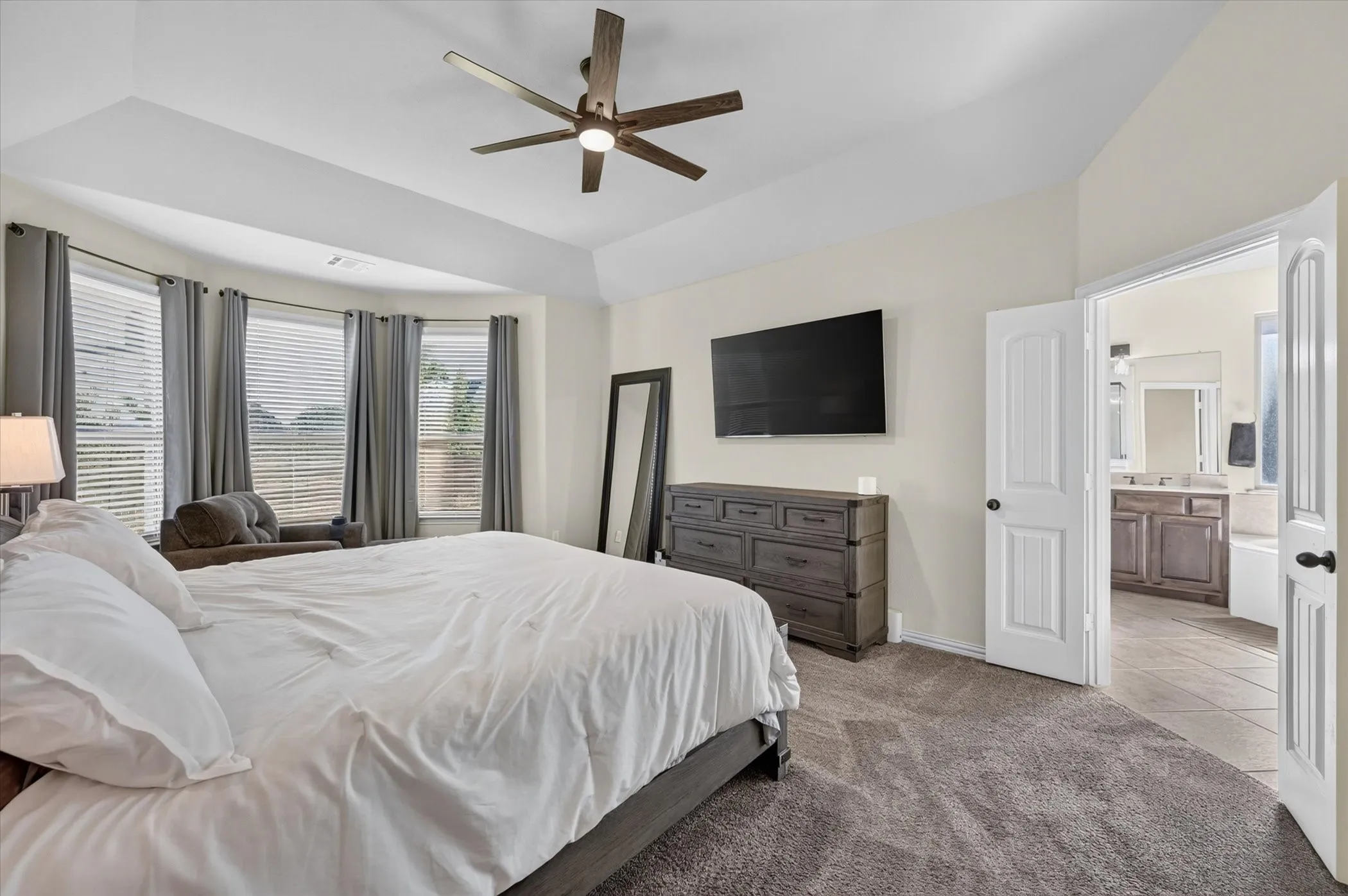 Spacious primary bedroom featuring a curved bay window arrangement with plantation shutters, ceiling fan with brushed metal finish, wall-mounted flat-screen television, dark wood dresser with mirror, carpeted flooring, and ensuite bathroom access through white painted doors