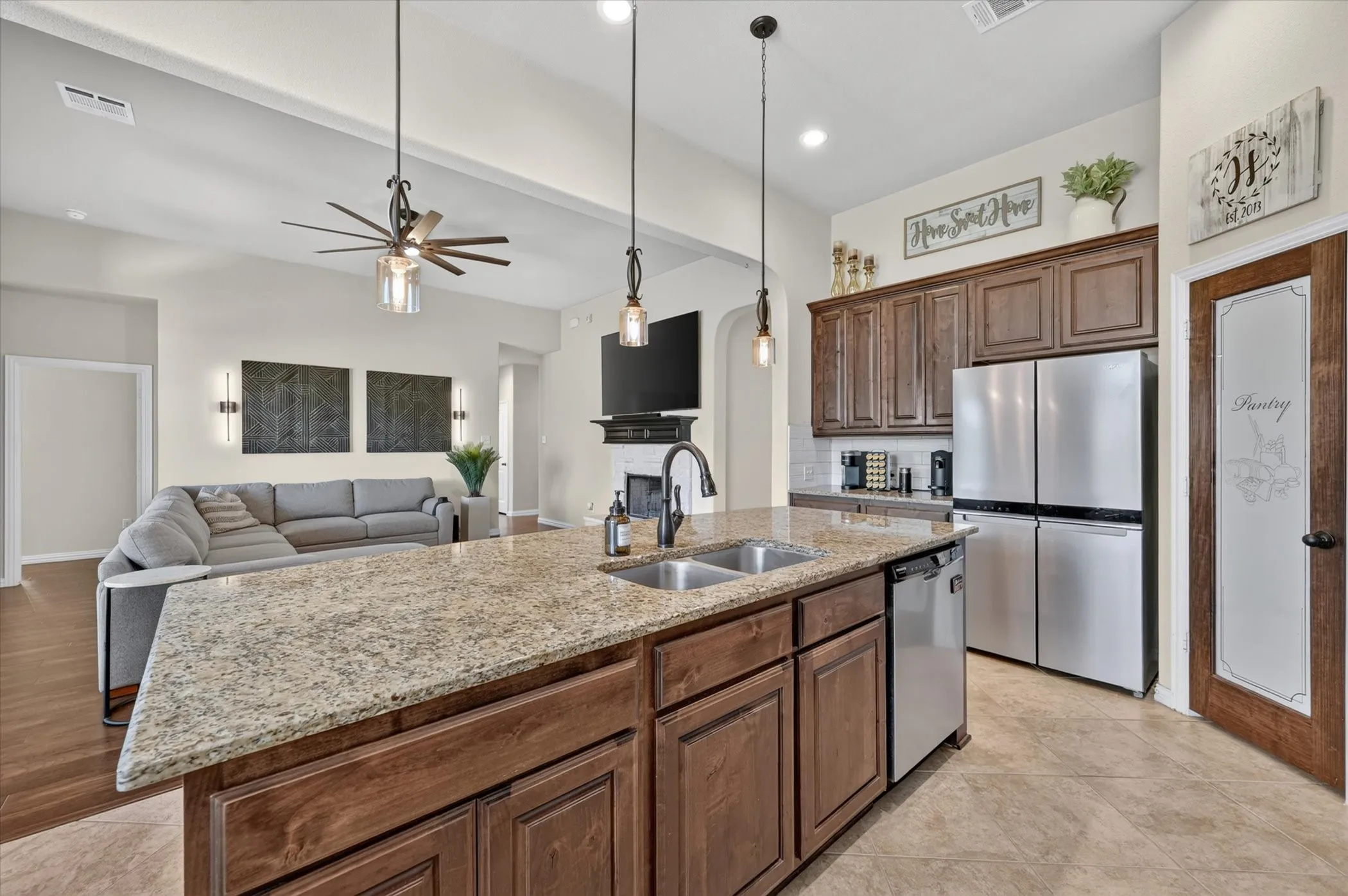 Open-concept kitchen and living area featuring a large granite-topped island with wood cabinetry, stainless steel appliances, pendant lighting, ceiling fans, and hardwood flooring throughout