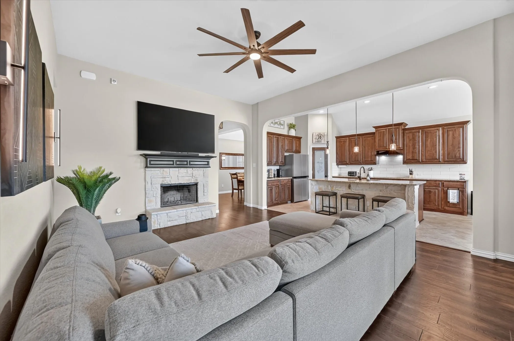 Vaulted ceiling with a ceiling fan, stone-clad fireplace, wood-finish flooring, and a kitchen featuring wood cabinetry and pendant lighting