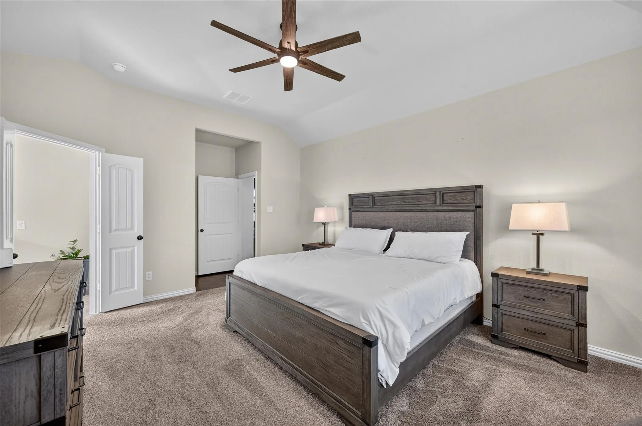Spacious primary bedroom with dark wood-finish bed frame and matching nightstands, neutral wall finishes, plush carpeting throughout, ceiling fan with integrated lighting, and dual closet access via white panel doors
