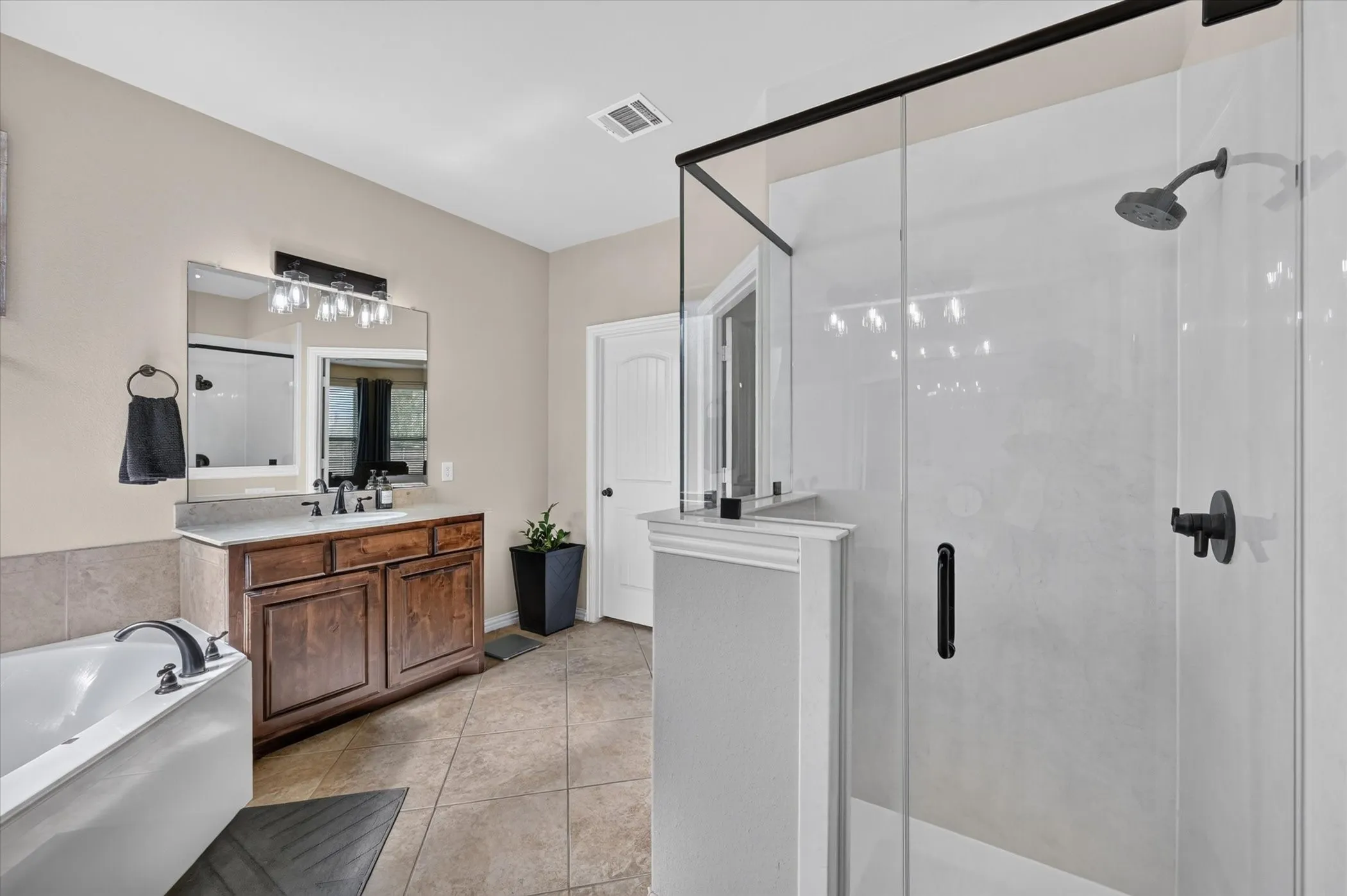 Spa-inspired primary bathroom featuring a freestanding soaking tub, a frameless glass-enclosed shower with tile surround and rainfall showerhead, dual-sink vanity with wood cabinetry and quartz countertop, and neutral tile flooring throughout