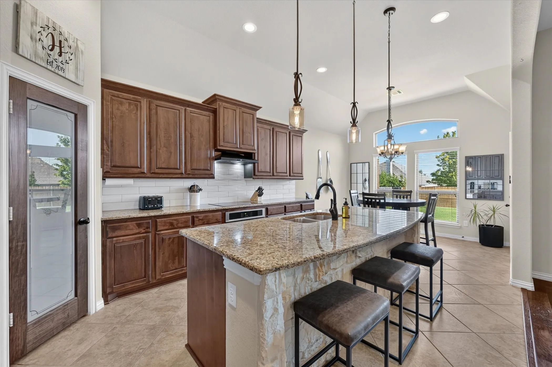 Chef's kitchen featuring rich wood cabinetry, granite-topped island with seating for three, stainless steel appliances, pendant lighting, and tile flooring that extends into the adjoining dining and living areas flooded with natural light through expansive windows
