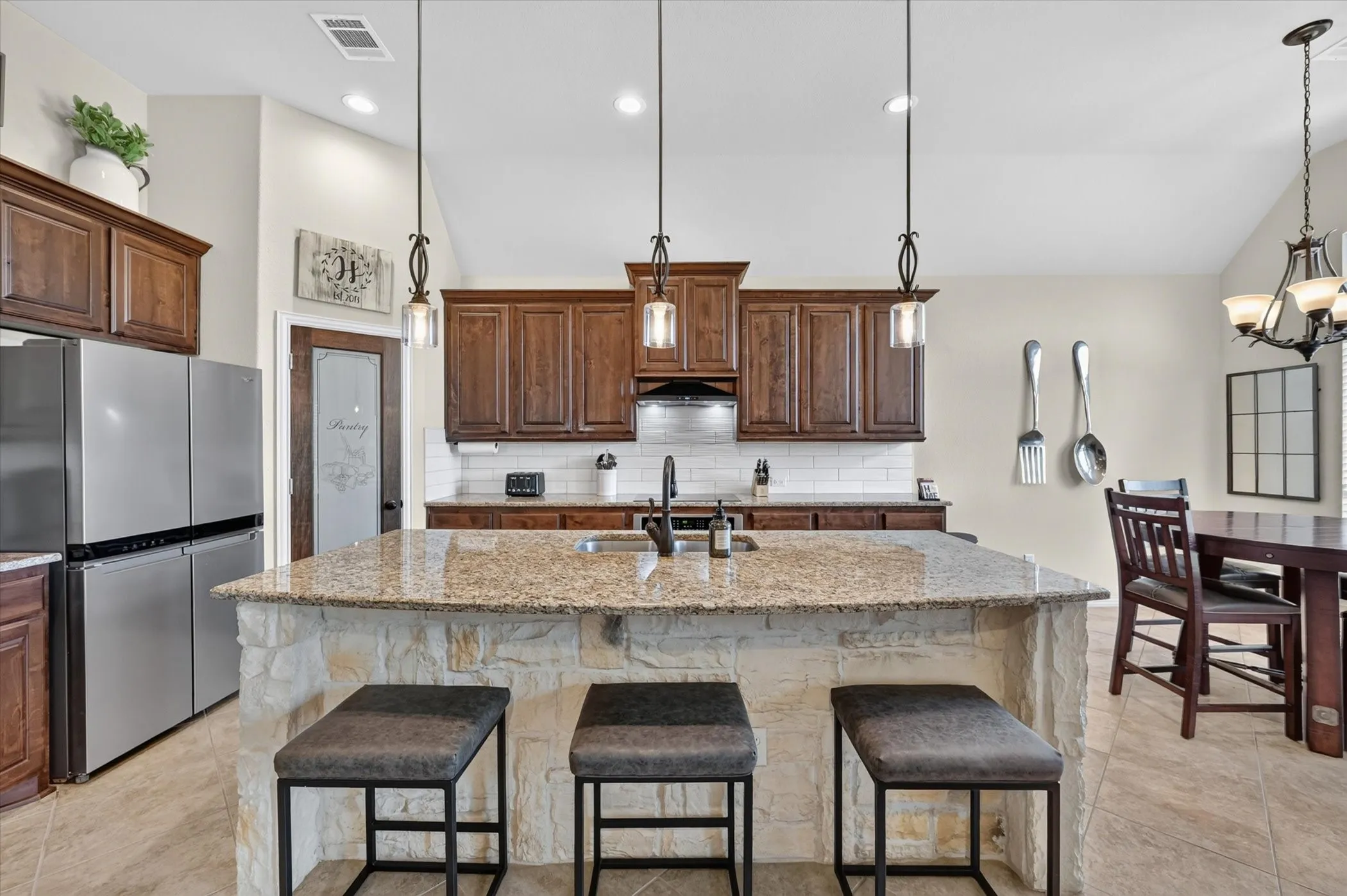 Chef's kitchen showcasing rich wood cabinetry, a large stone-topped island with seating for three, pendant lighting, stainless steel appliances, and a coordinating dining area with hardwood-finish flooring throughout