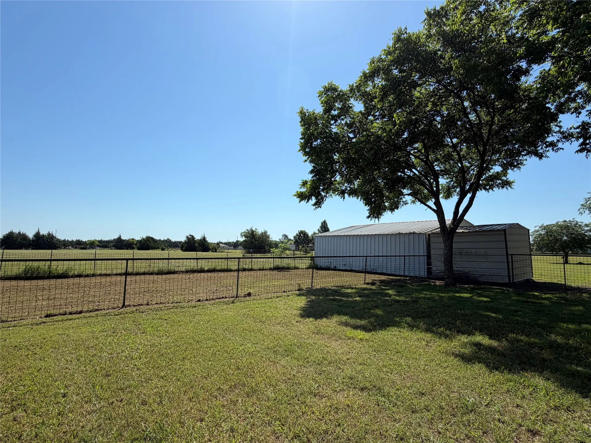 Expansive grassy terrain featuring a metal outbuilding with a metal roof, mature shade tree, and perimeter fencing