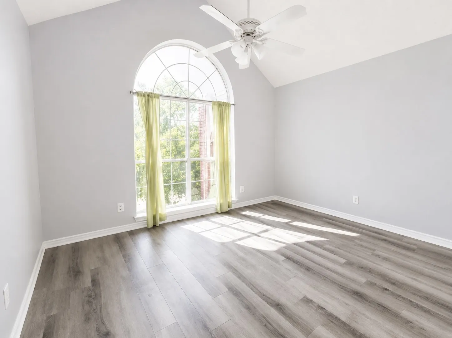 Bright, sun-filled room showcasing an arched transom window with natural light, neutral wall finishes, wood-finish flooring with gray tones, ceiling fan, and multiple electrical outlets for versatile furnishing options