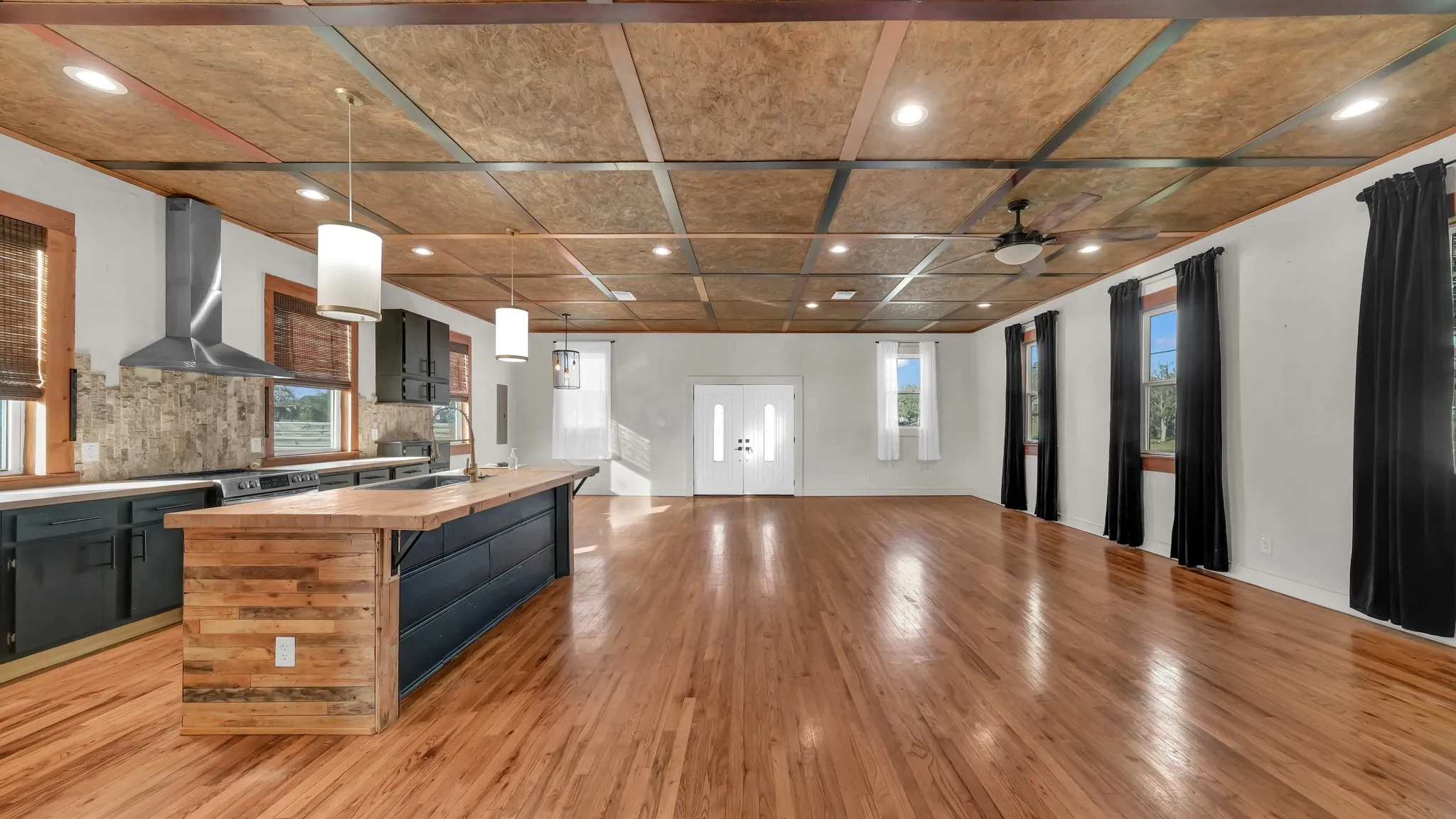 Hardwood flooring, wood-panel island with a sink, pendant lighting, stainless steel range hood, and double entry doors