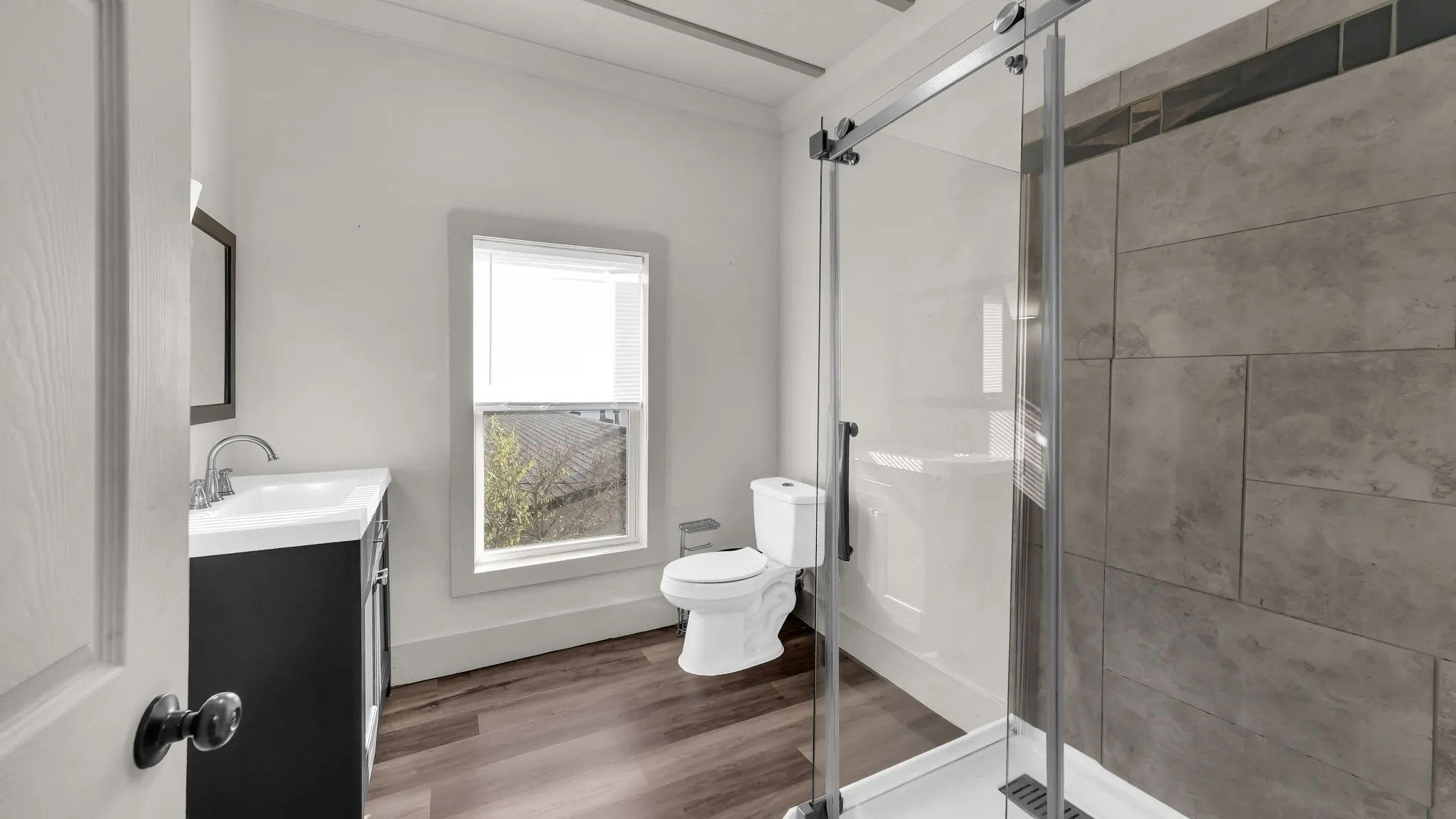 Modern primary bathroom featuring a frameless glass shower enclosure with gray tile surround, white subway tile accent wall, floating vanity with integrated sink and matte black hardware, low-flow toilet, wood-finish flooring, and natural light from an operable window