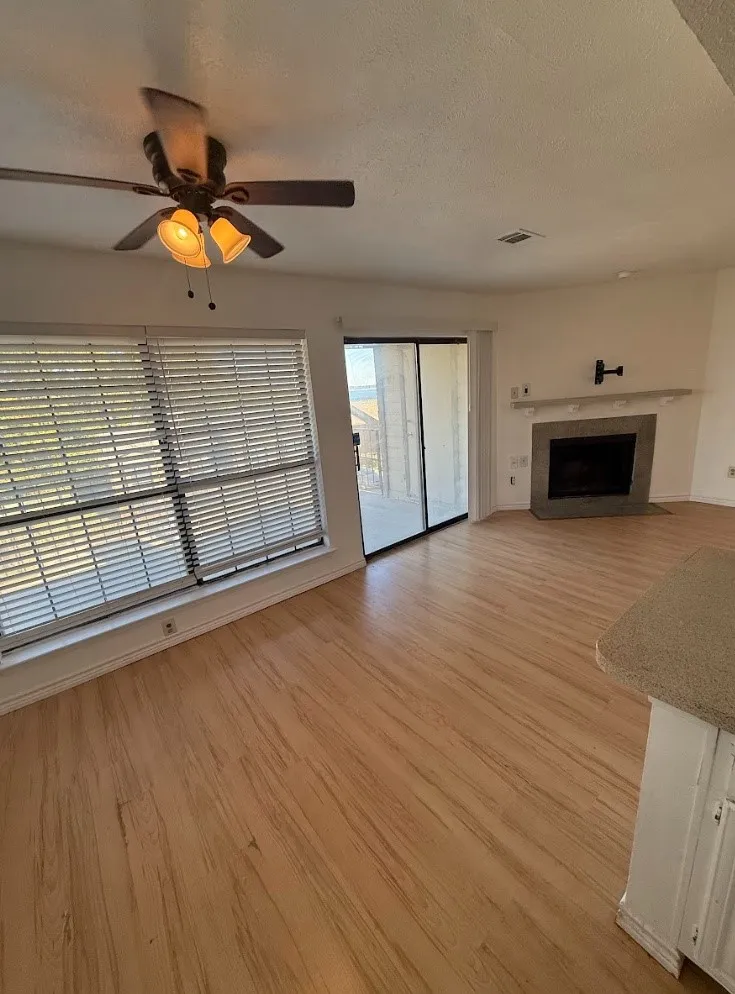 Wood-finish flooring, ceiling fan with integrated lighting, fireplace with stone surround and mantel, sliding glass door, and large window with blinds