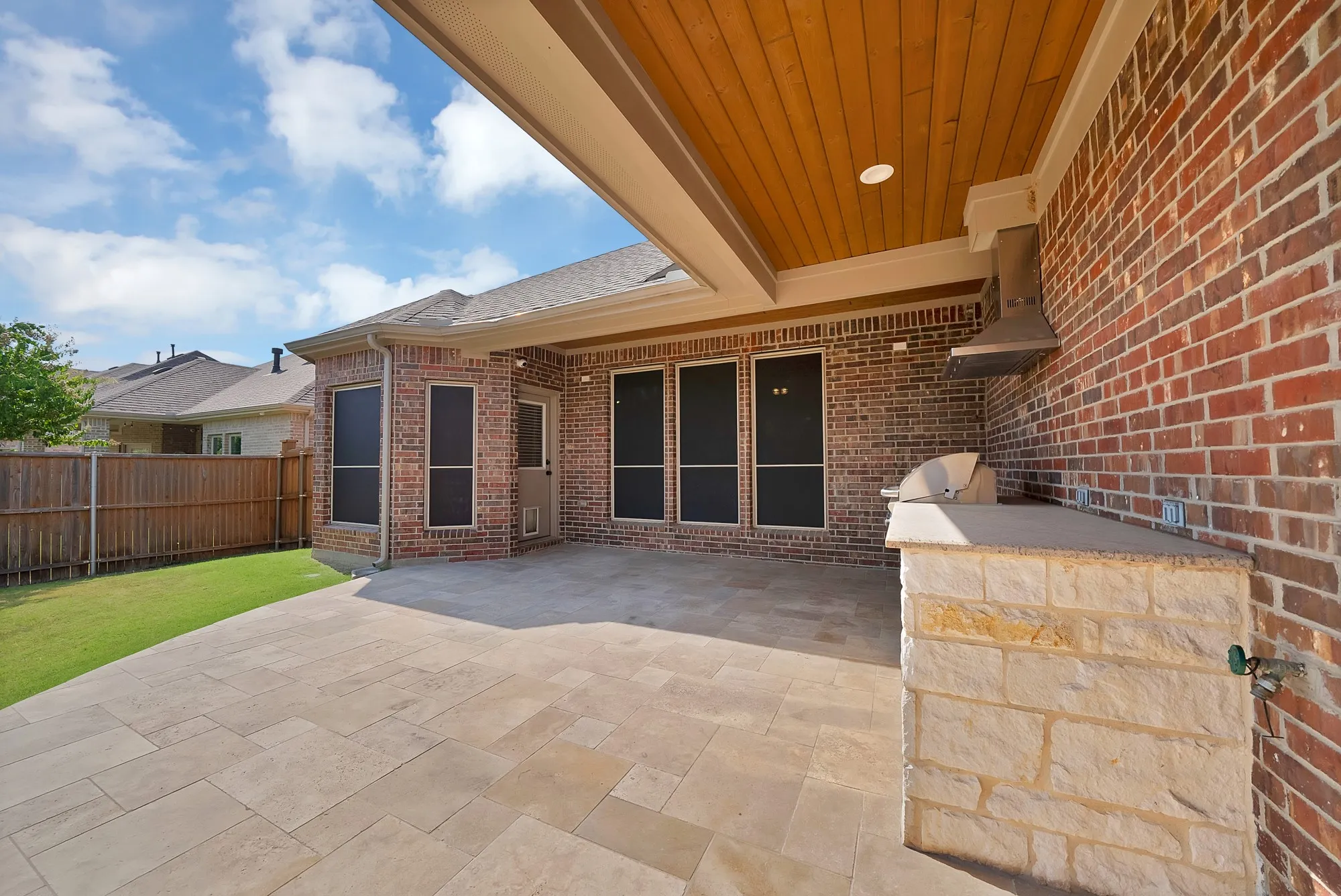Expansive covered patio with a wood-plank ceiling, built-in outdoor kitchen with a grill and stone-clad base, large format tile flooring, and a fenced yard