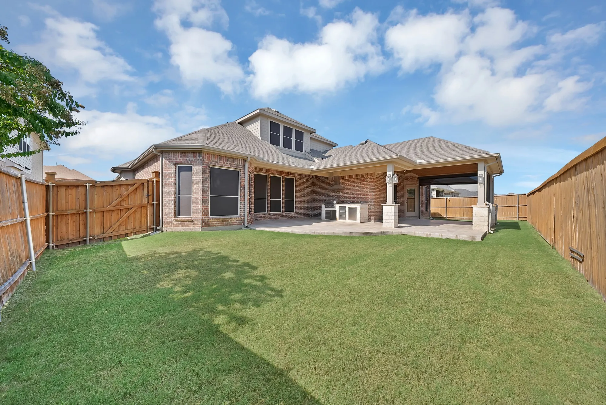 Expansive backyard with a manicured lawn, brick exterior, covered patio, stone pillars, and wood fencing