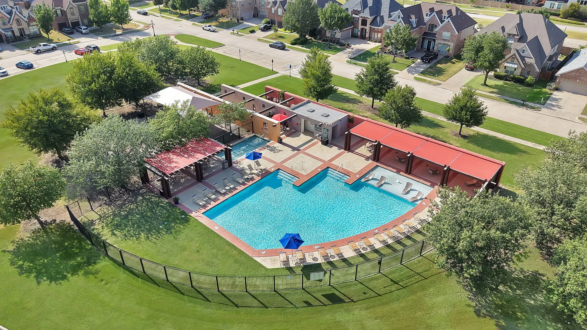Expansive community pool with a surrounding stone-finish deck, multiple covered lounge areas, and a black wrought-iron perimeter fence