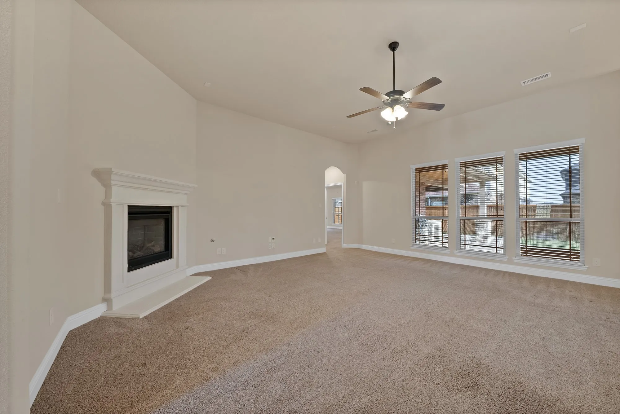 Vaulted ceiling with a ceiling fan and light fixture, three large windows with blinds, a built-in fireplace with a white mantel, and neutral-toned carpeting