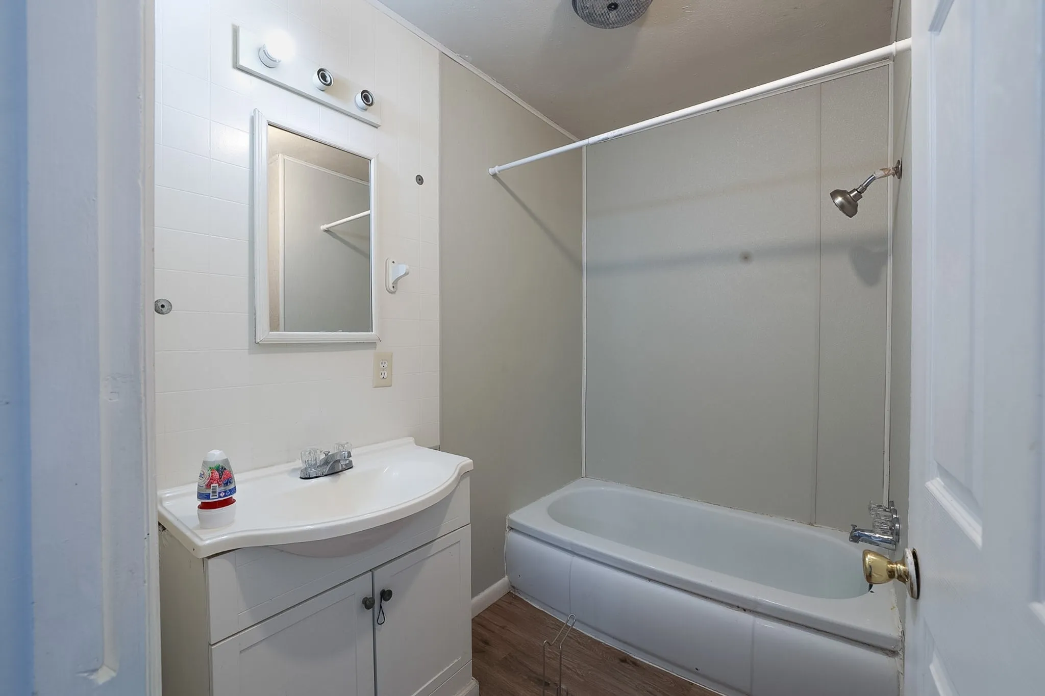 Updated bathroom featuring a curved vanity with white cabinetry, an illuminated mirror with integrated lighting, a tile-surround bathtub with glass enclosure and chrome fixtures, and wood-finish flooring