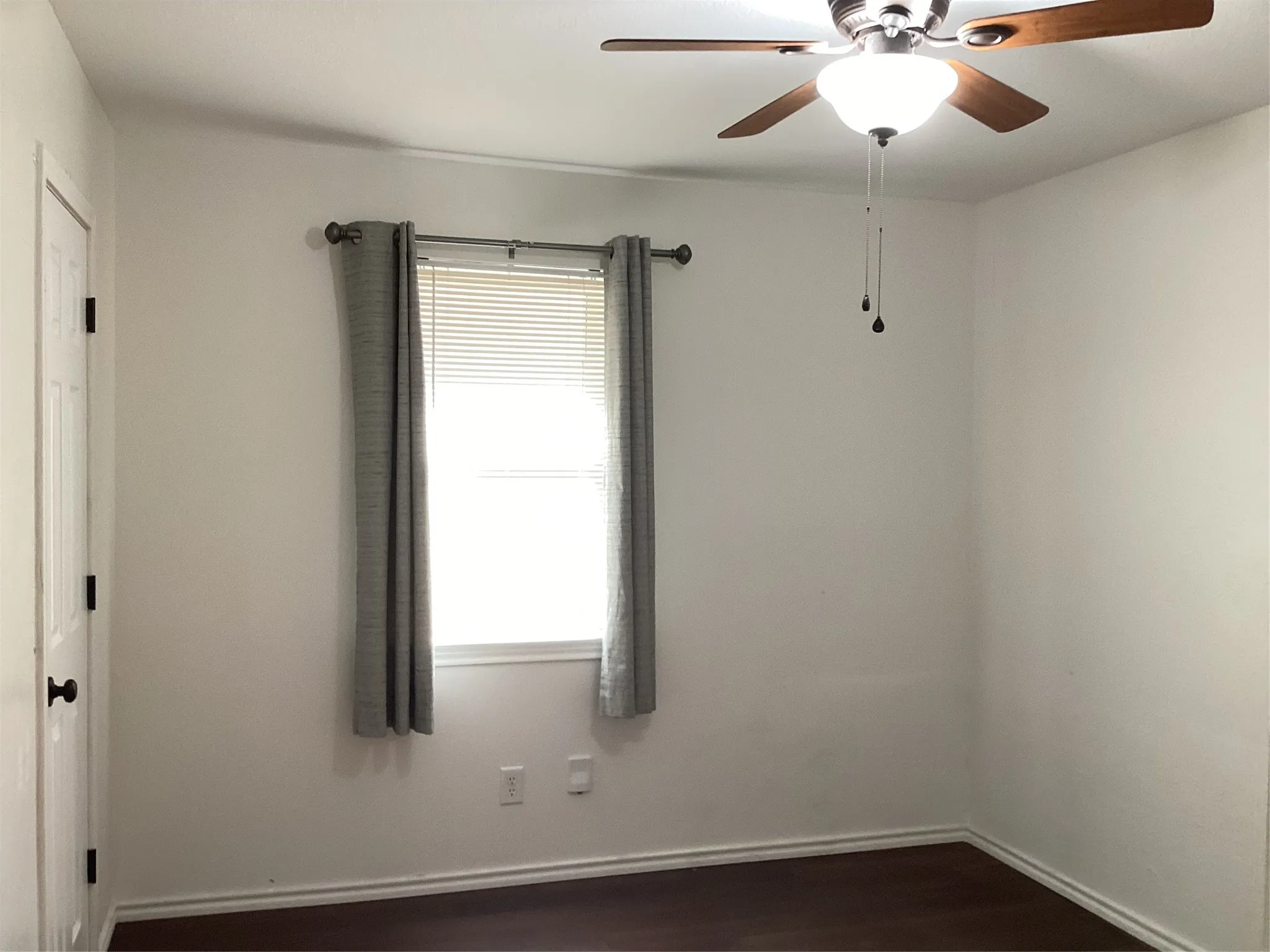 Wood-finish flooring with white baseboards, window with blinds, and a ceiling fan with light fixture
