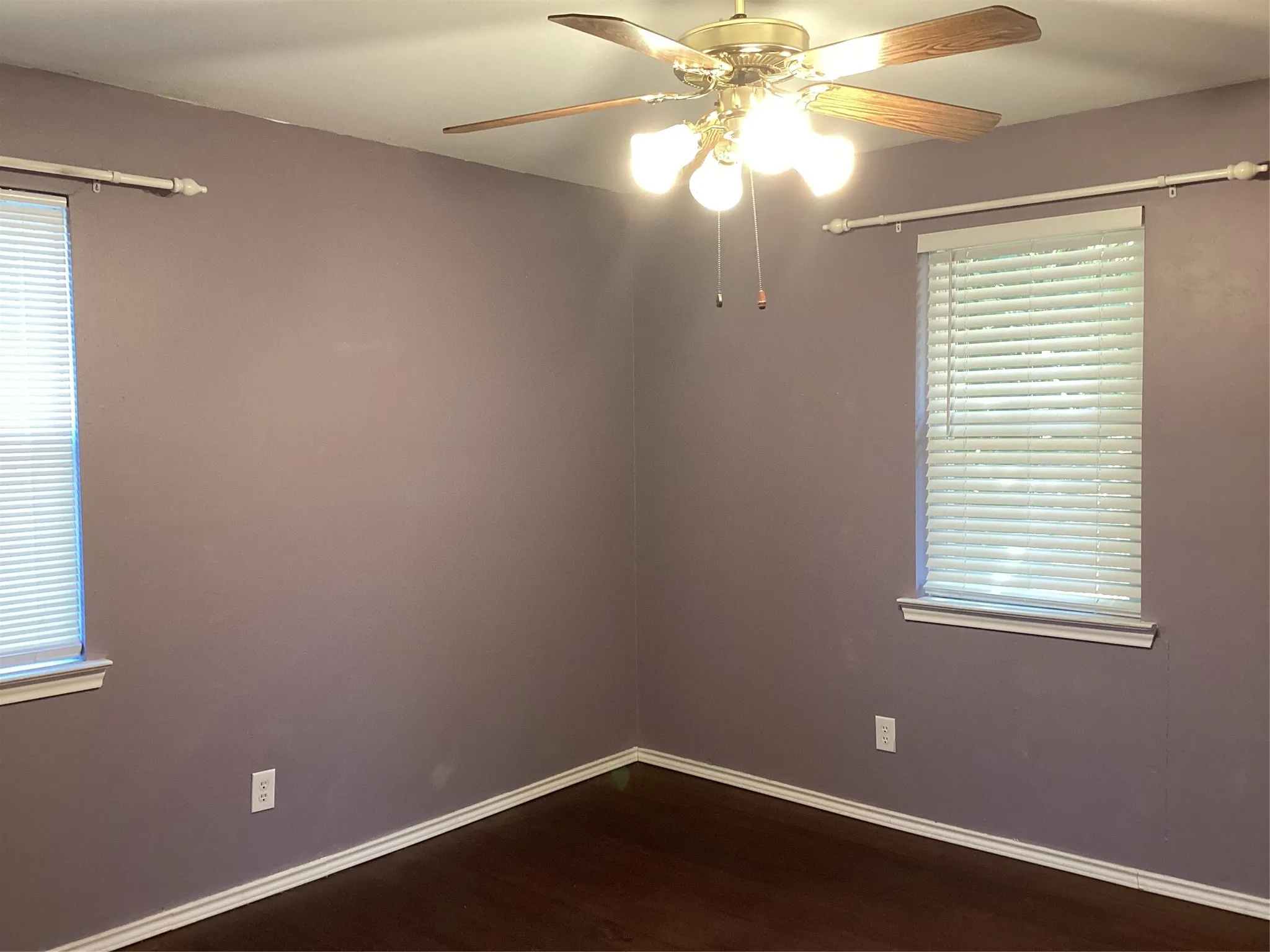 Room featuring wood-finish flooring, two windows with blinds, a ceiling fan with light fixture, and white trim