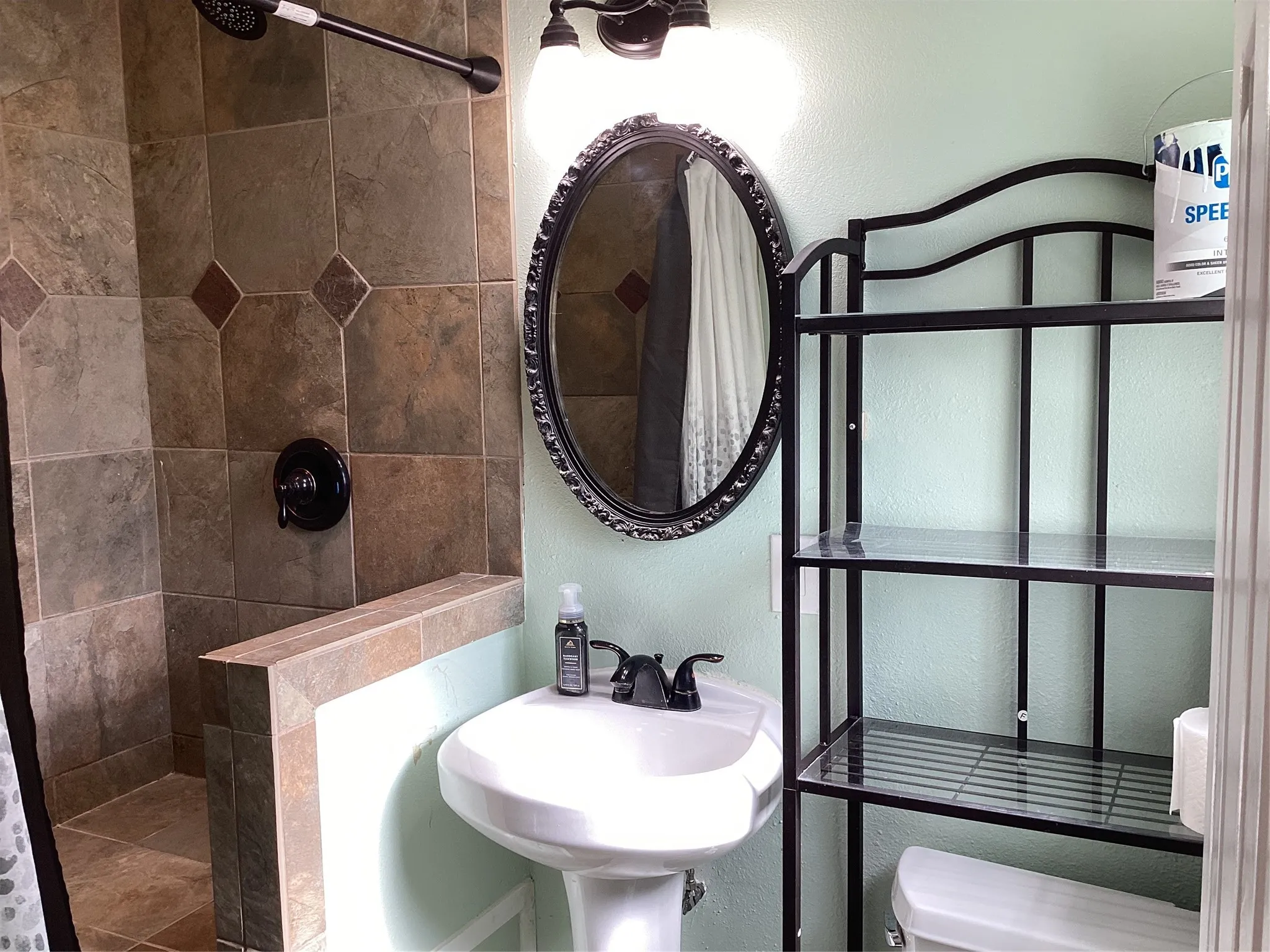 Tile shower with dark bronze fixtures, pedestal sink with dark bronze faucet, and an oval vanity mirror with an ornate frame