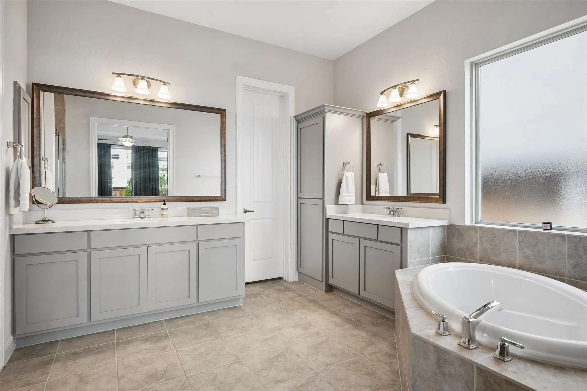 Spa-inspired primary bathroom featuring dual vanities with gray cabinetry and framed mirrors, stone-finish flooring throughout, a soaking tub with chrome fixtures, and abundant natural light.