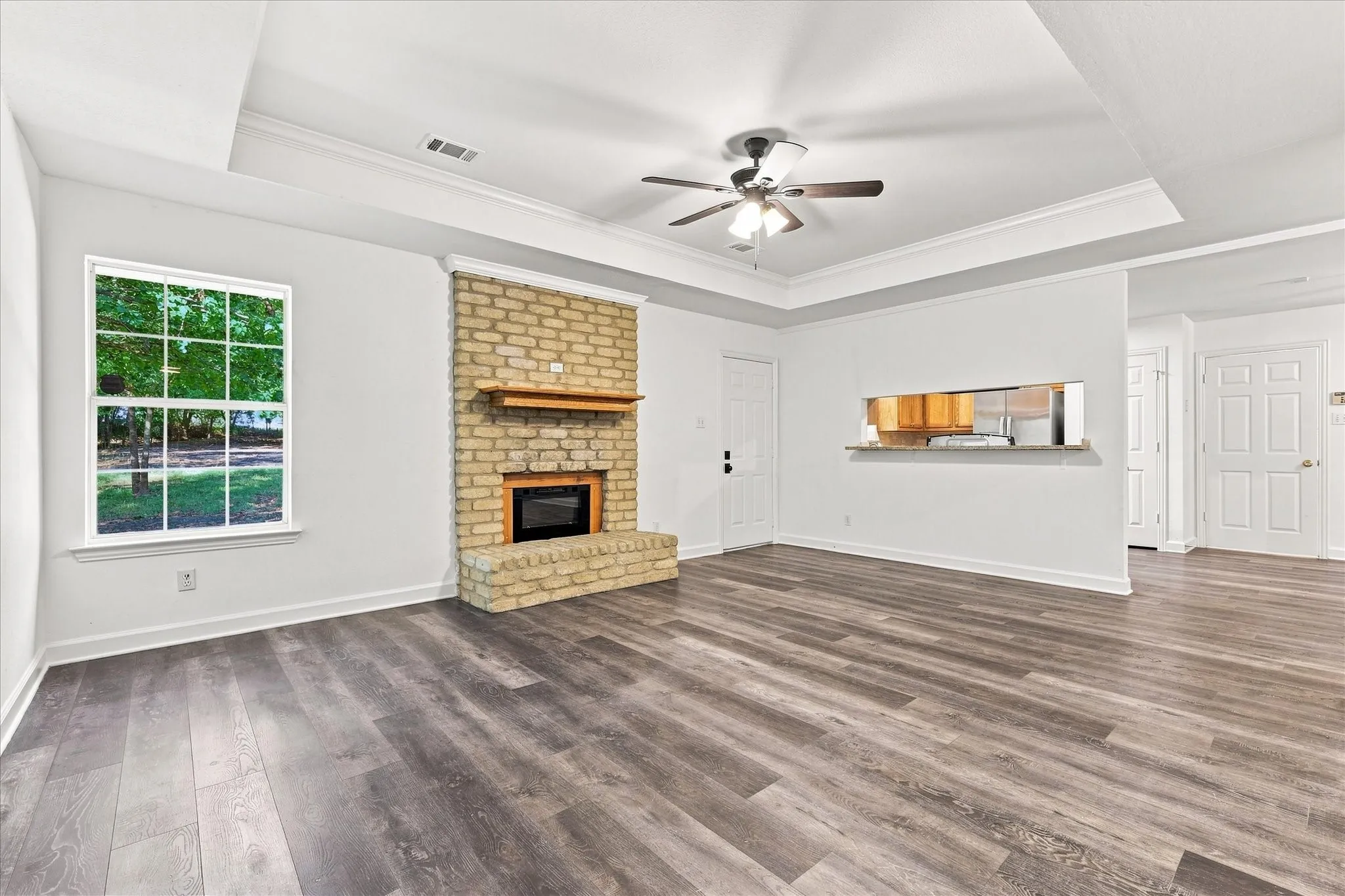 Tray ceiling with crown molding, wood-finish flooring, brick fireplace with wood mantel, ceiling fan, and pass-through window with counter
