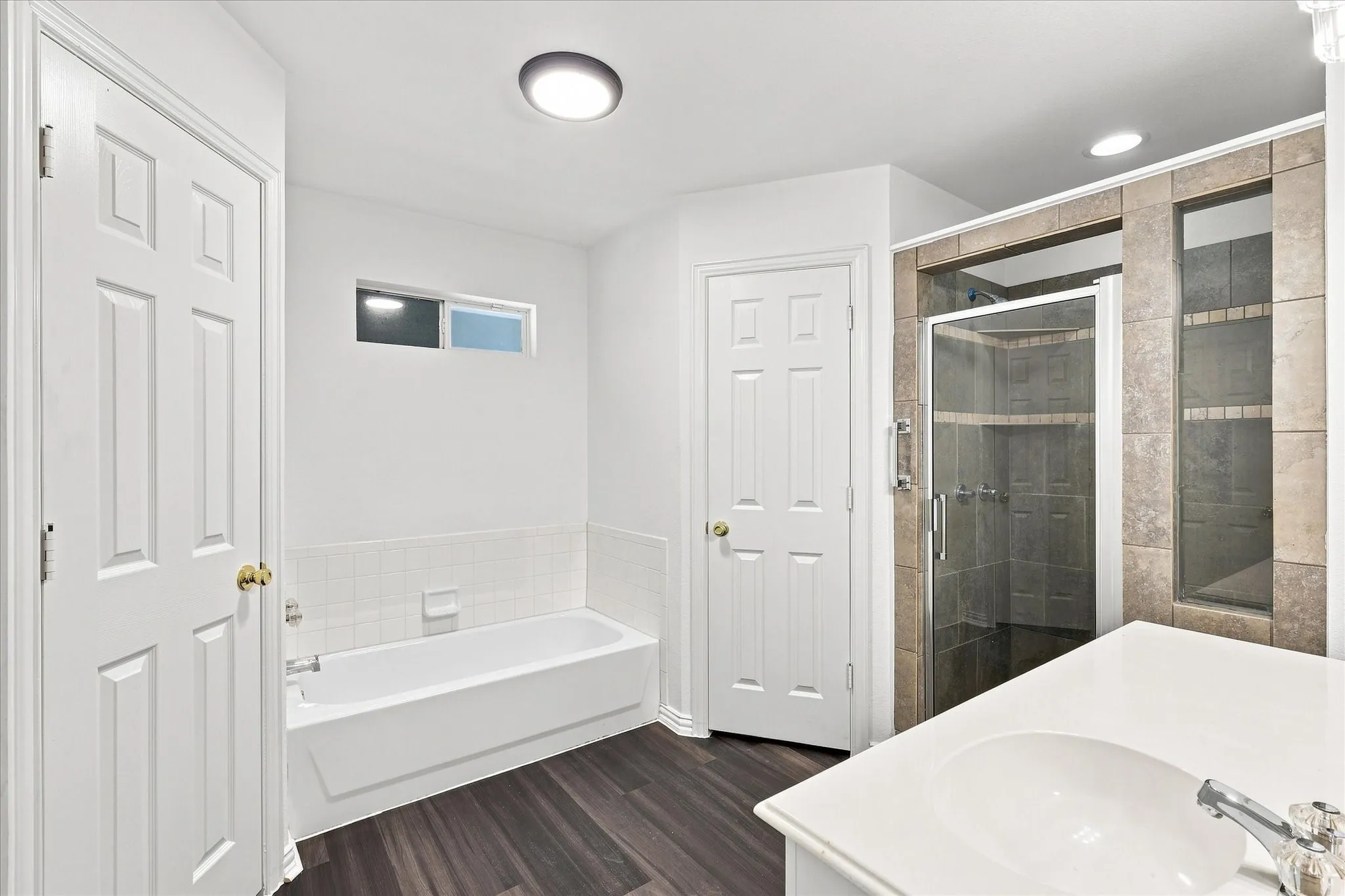 Bathroom featuring a white bathtub with subway tile surround, wood-finish flooring, a glass-enclosed shower with tile, and a vanity with an integrated sink