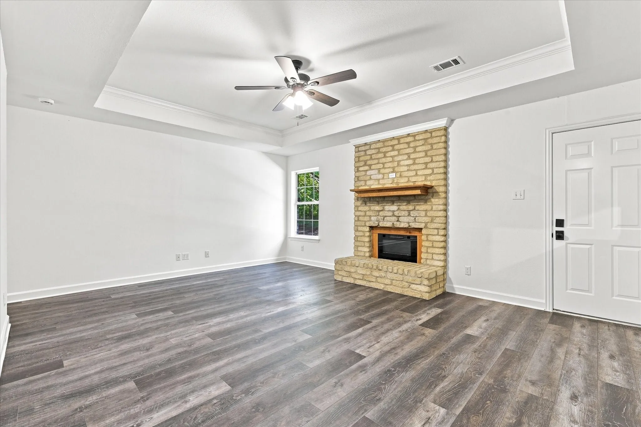 Tray ceiling with crown molding, wood-finish flooring, brick fireplace with wood mantel, ceiling fan, and paneled door