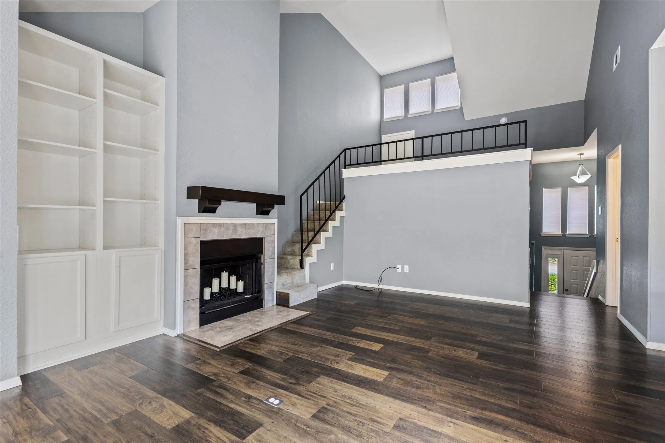 Living area featuring wood-finish flooring, a fireplace with a stone surround, built-in shelving, a staircase with a metal railing, and high ceilings
