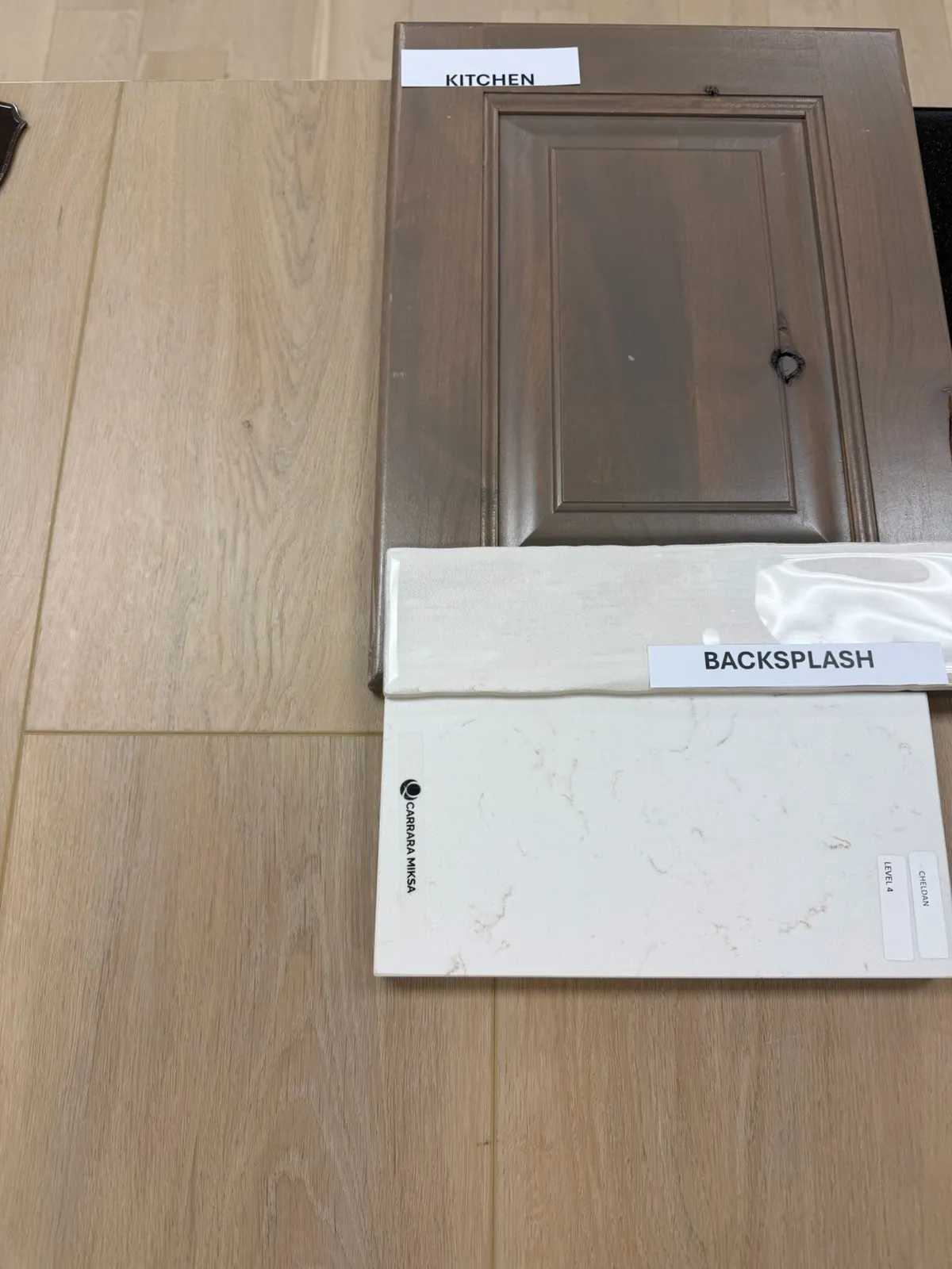 Wood-finish flooring with a light tone, a brown paneled cabinet door, a light-colored backsplash sample, and a white countertop sample with subtle veining