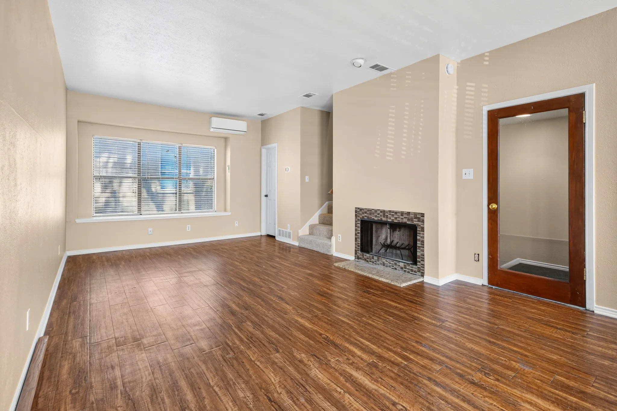 Hardwood-finish flooring, a contemporary fireplace with a stone tile surround, a large window with blinds, a split-unit air conditioner, and a wood-framed glass door