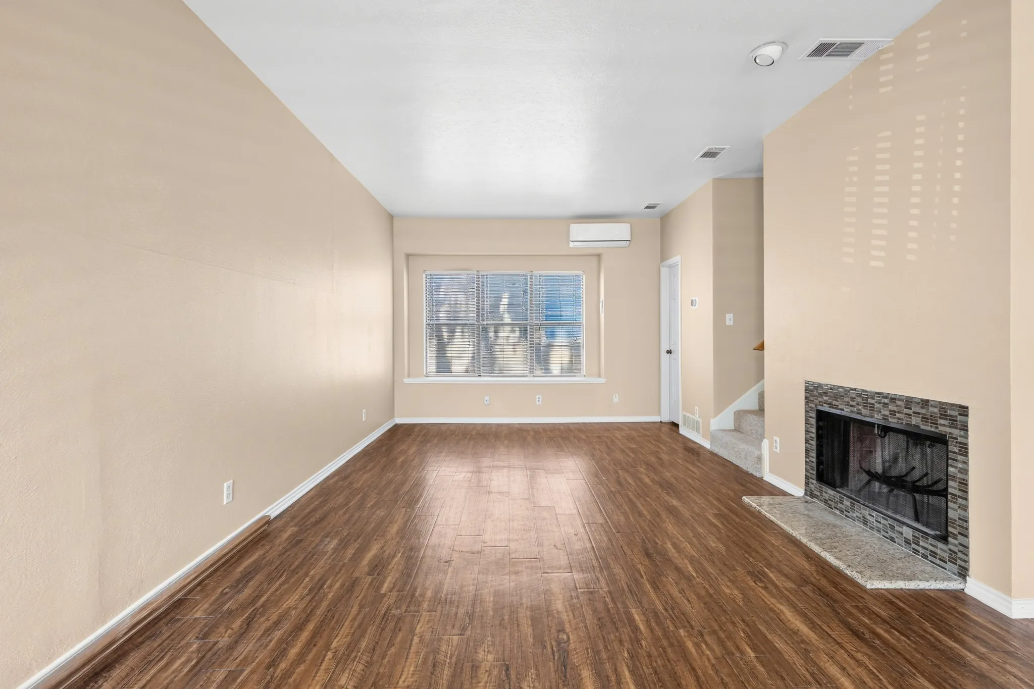 Wood-finish flooring, accent wall with fireplace featuring dark tile surround, light-colored walls, window with blinds, and mini-split air conditioning unit