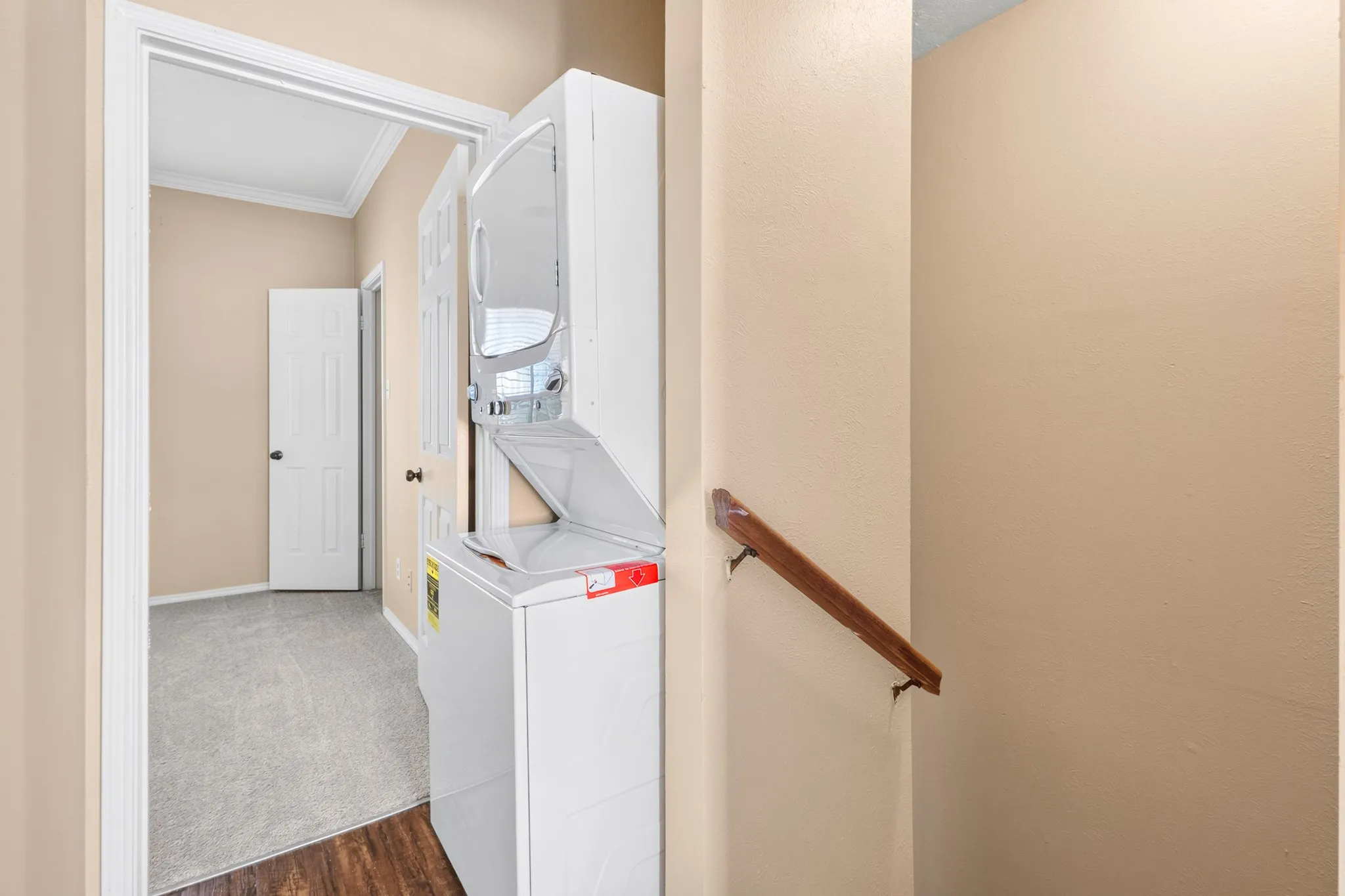 Stacked washer and dryer unit, wood-finish flooring, light-toned wall paint, white trim, and a wooden handrail