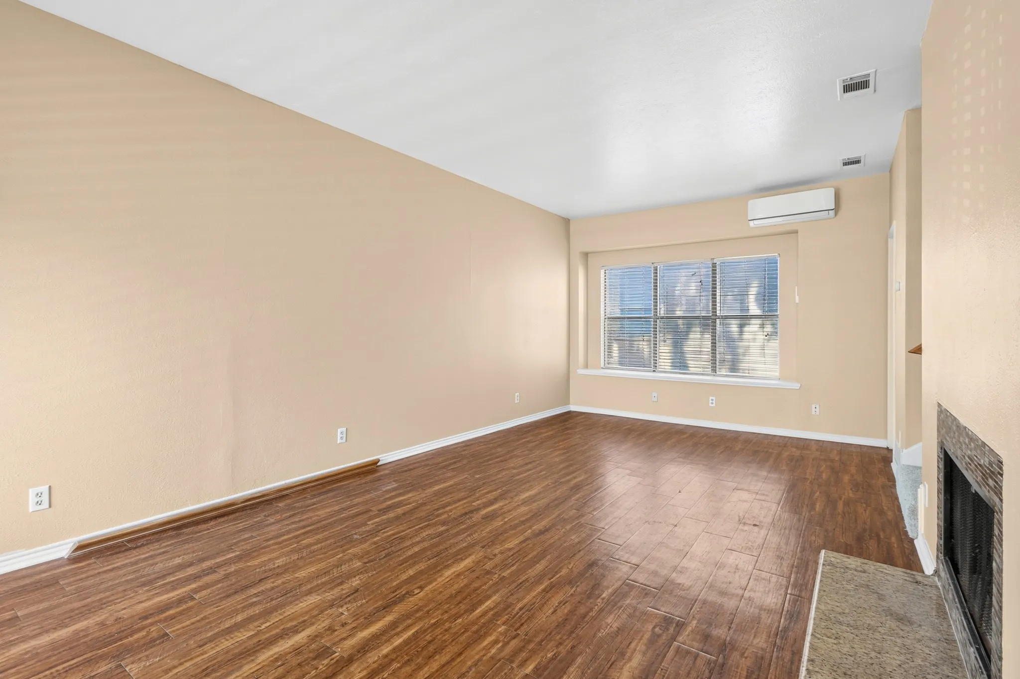 Wood-finish flooring, multiple windows with blinds, built-in fireplace with hearth, and wall-mounted climate control unit
