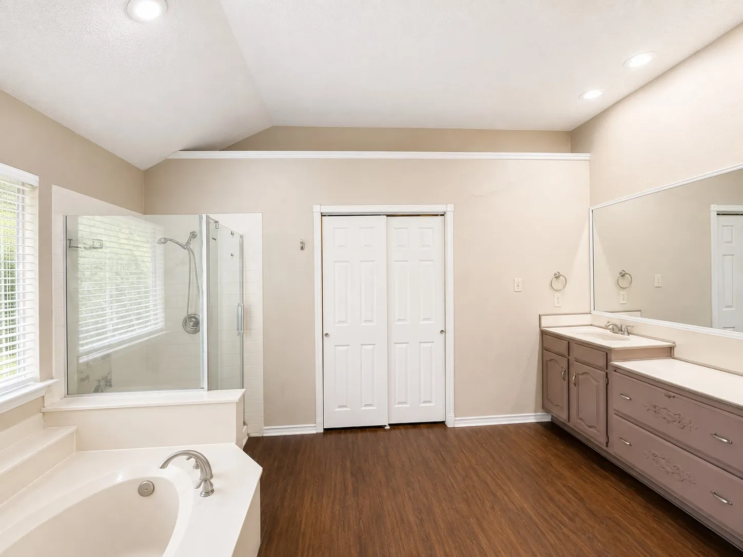 Vaulted ceiling with recessed lighting, wood-finish flooring, built-in soaking tub, glass-enclosed shower, and dual vanity with integrated mirror