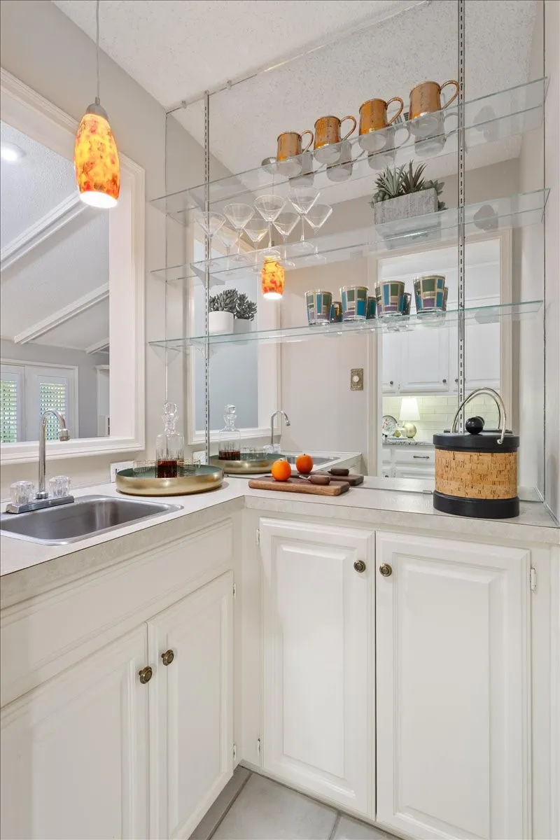 Corner wet bar with an integrated sink, white cabinetry, overhead glass shelving, and a pendant light fixture