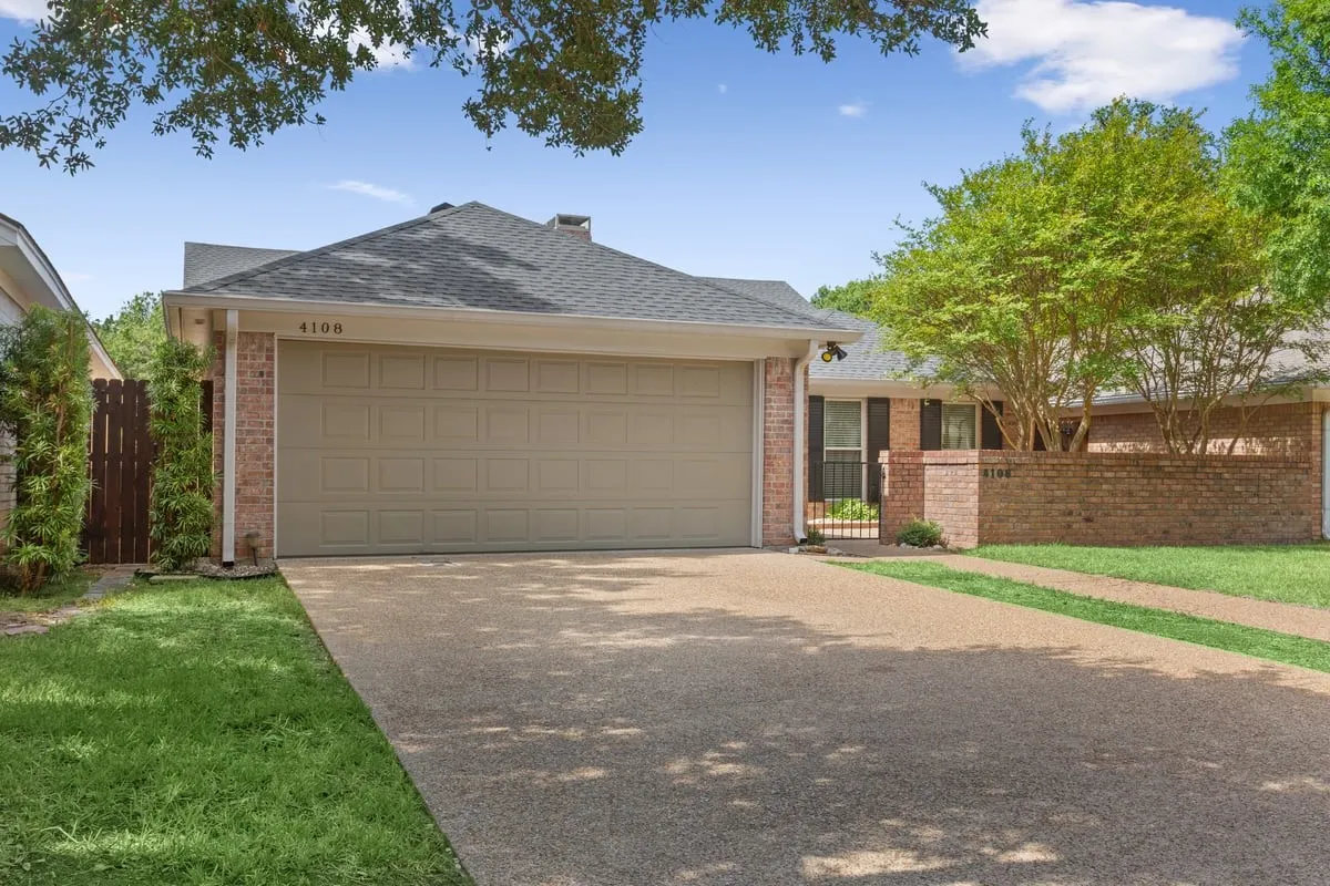 Brick exterior featuring a two-car garage, gravel driveway, and mature landscaping