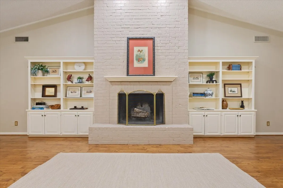 Vaulted ceiling, painted brick fireplace with hearth, brass-finish fire screen, built-in shelving, and hardwood flooring