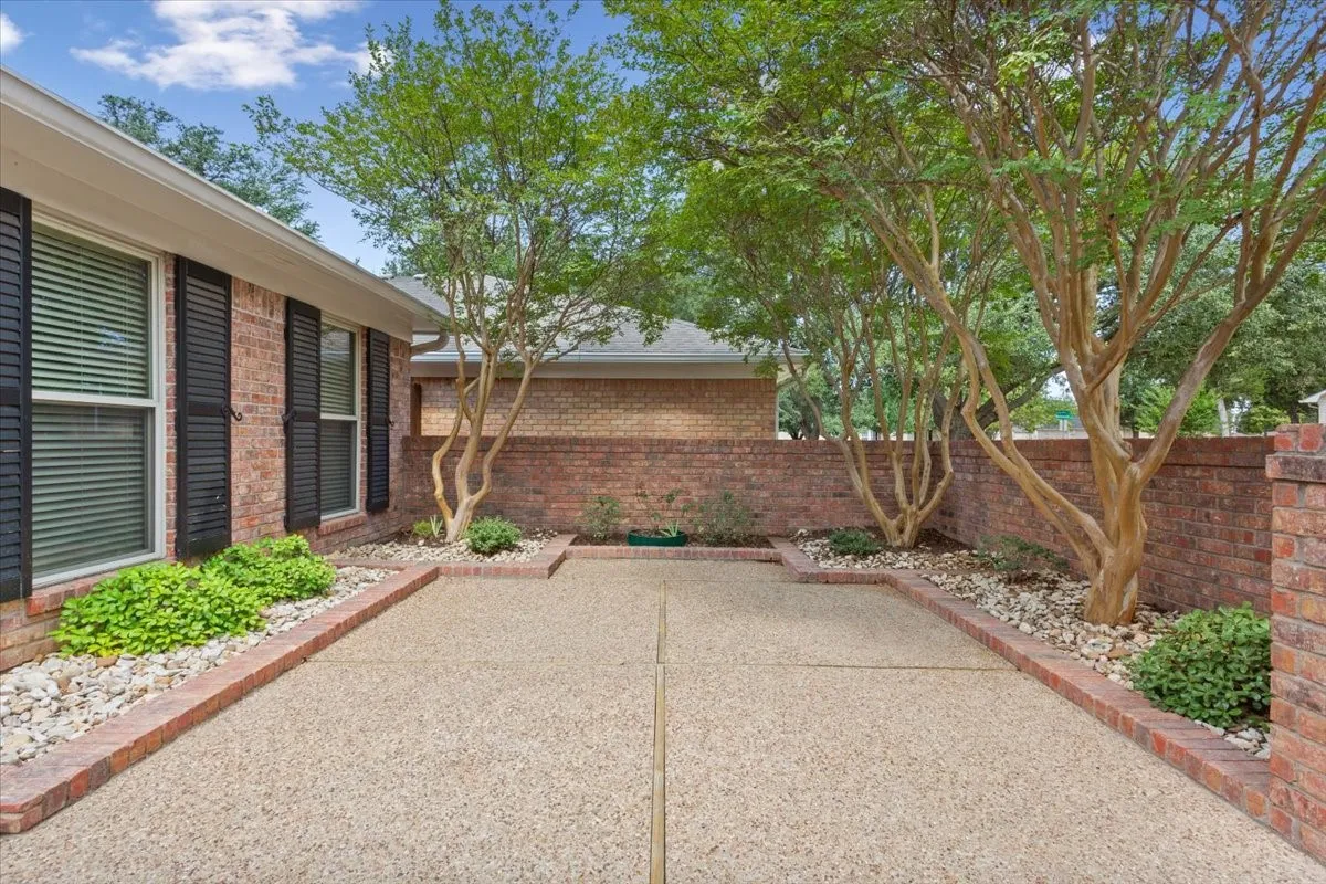 Brick residence with modern architectural details and a generous front courtyard featuring mature shade trees, decorative river rock landscaping, and bordered planting beds