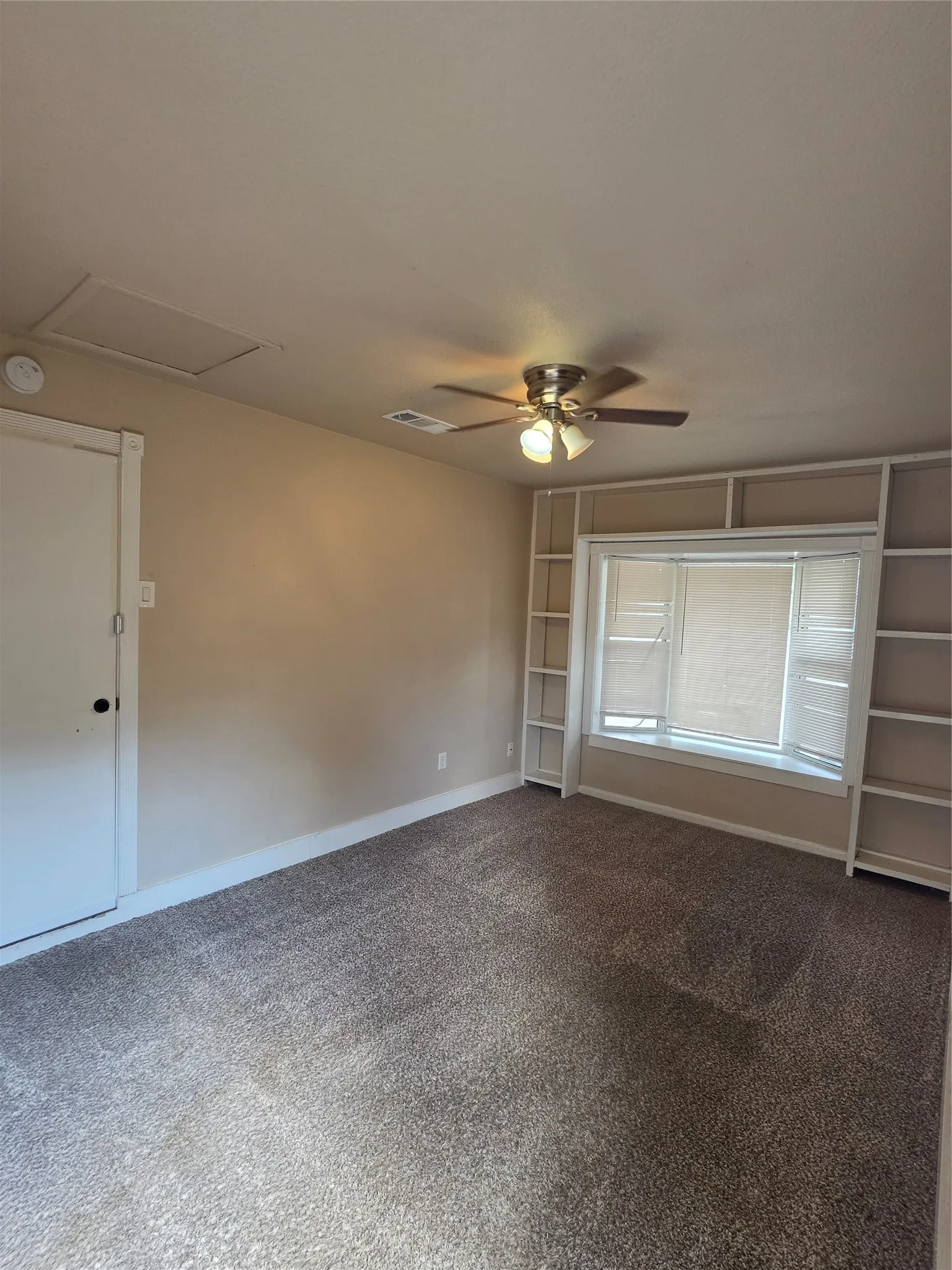 Carpeted room featuring a ceiling fan, built-in shelving, and a bay window with blinds