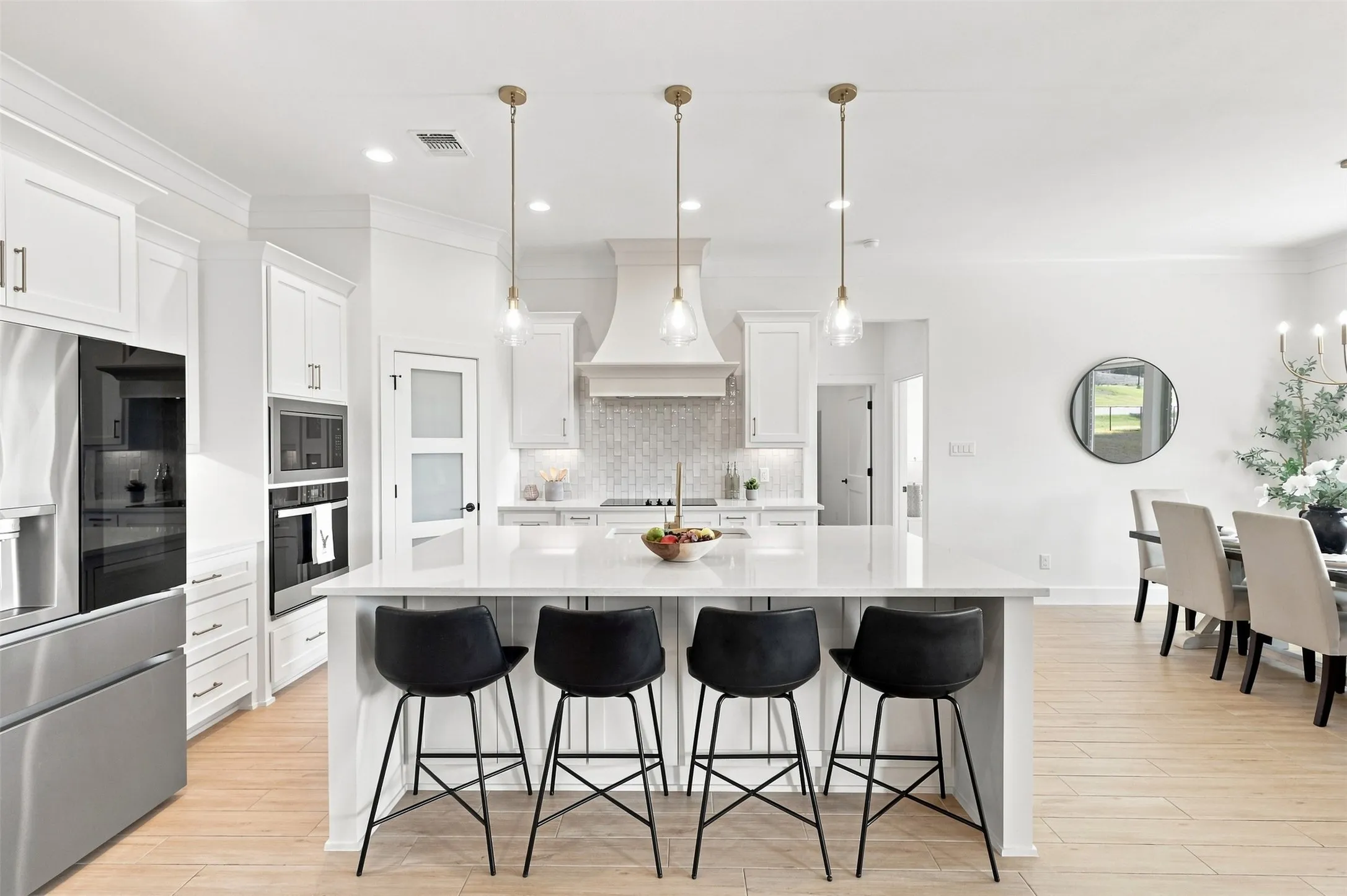 Open-concept kitchen and dining area featuring a large waterfall-edge island with seating for four, white cabinetry throughout, stainless steel appliances including built-in refrigerator and wall oven, pendant lighting above the island, wood-finish flooring, and seamless sightlines to the adjacent dining space