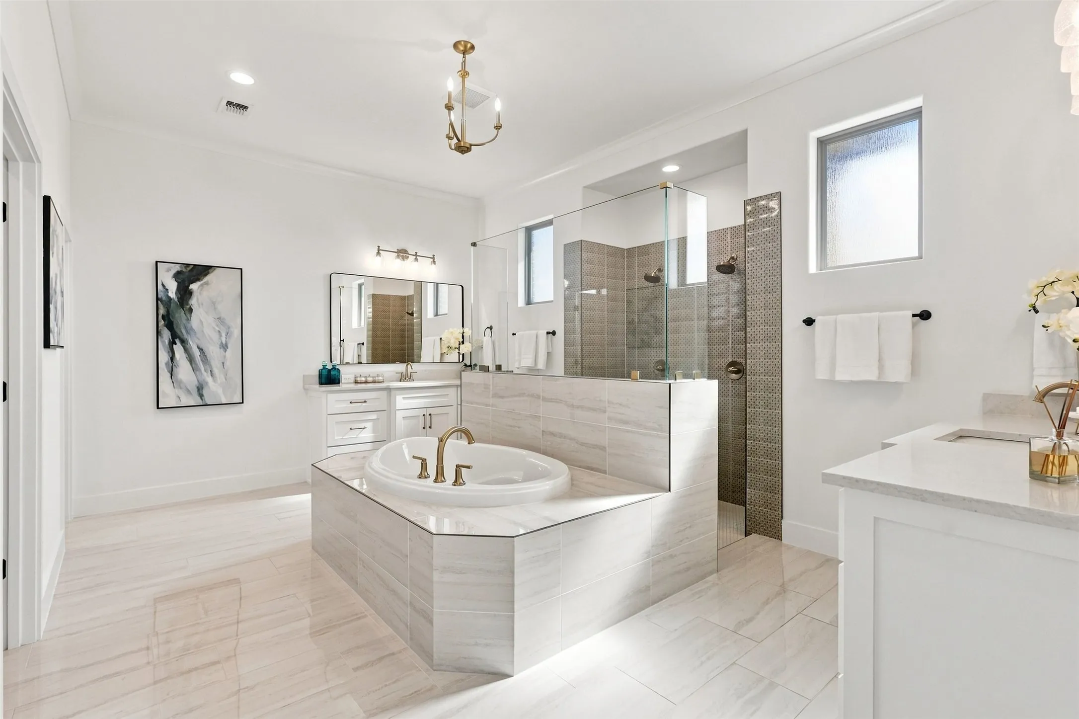 Spacious bathroom with light-toned tile flooring, a large soaking tub with brass fixtures, a glass-enclosed shower, dual vanity with white cabinetry, and a separate vanity with a single basin