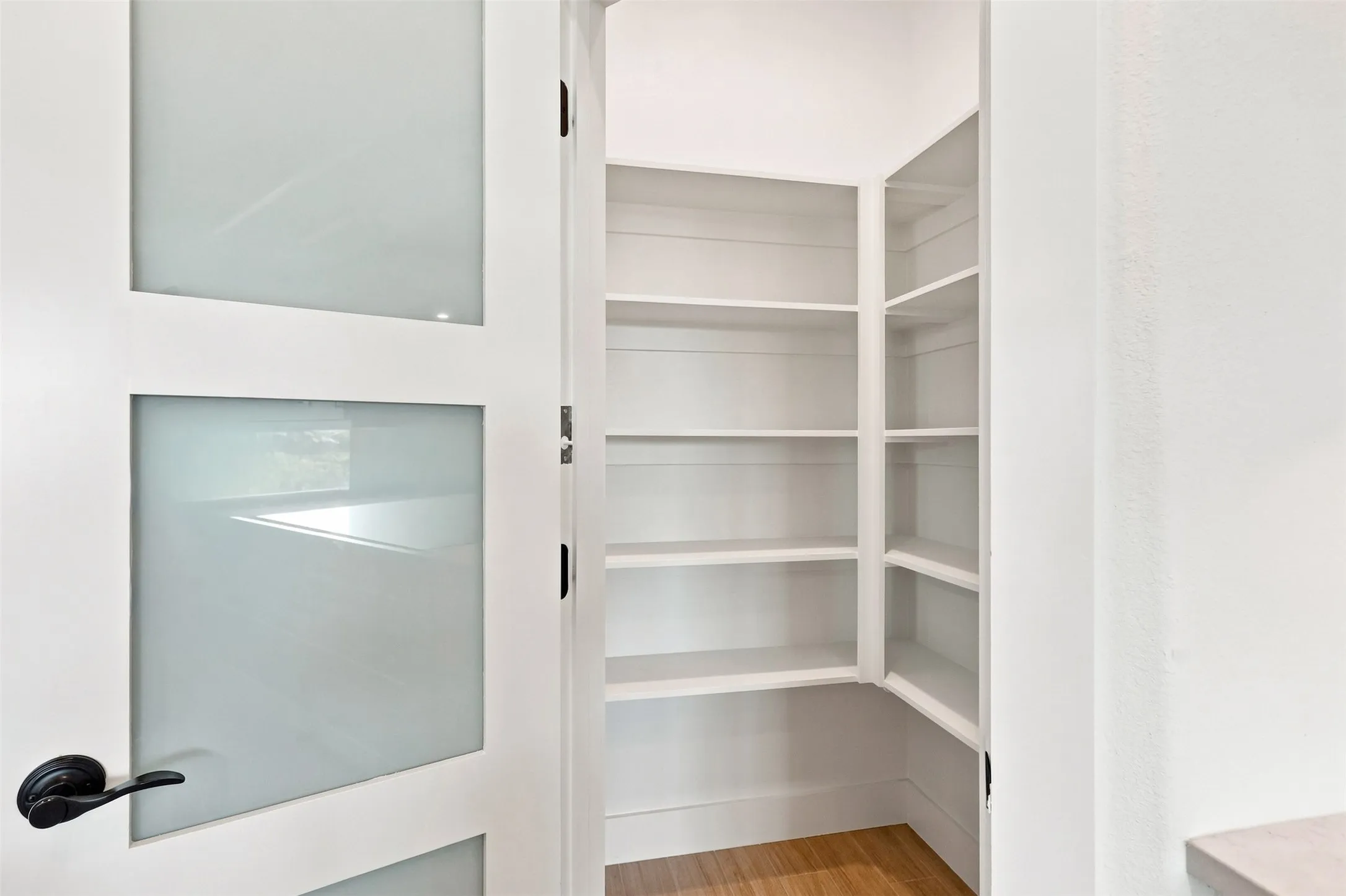 White door with frosted glass panels and black hardware, revealing extensive built-in shelving and wood-finish flooring and a built in hinge switch for lighting