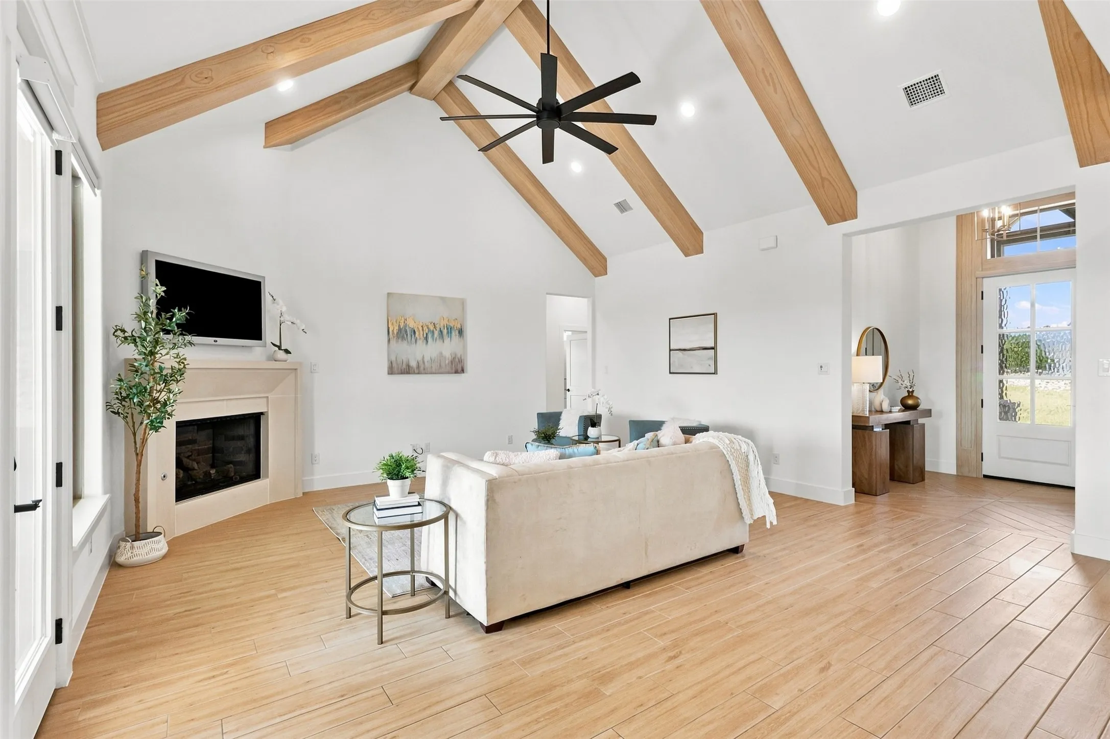 Vaulted ceiling with exposed wood beams, wood-finish flooring, corner fireplace with stone surround, modern ceiling fan.