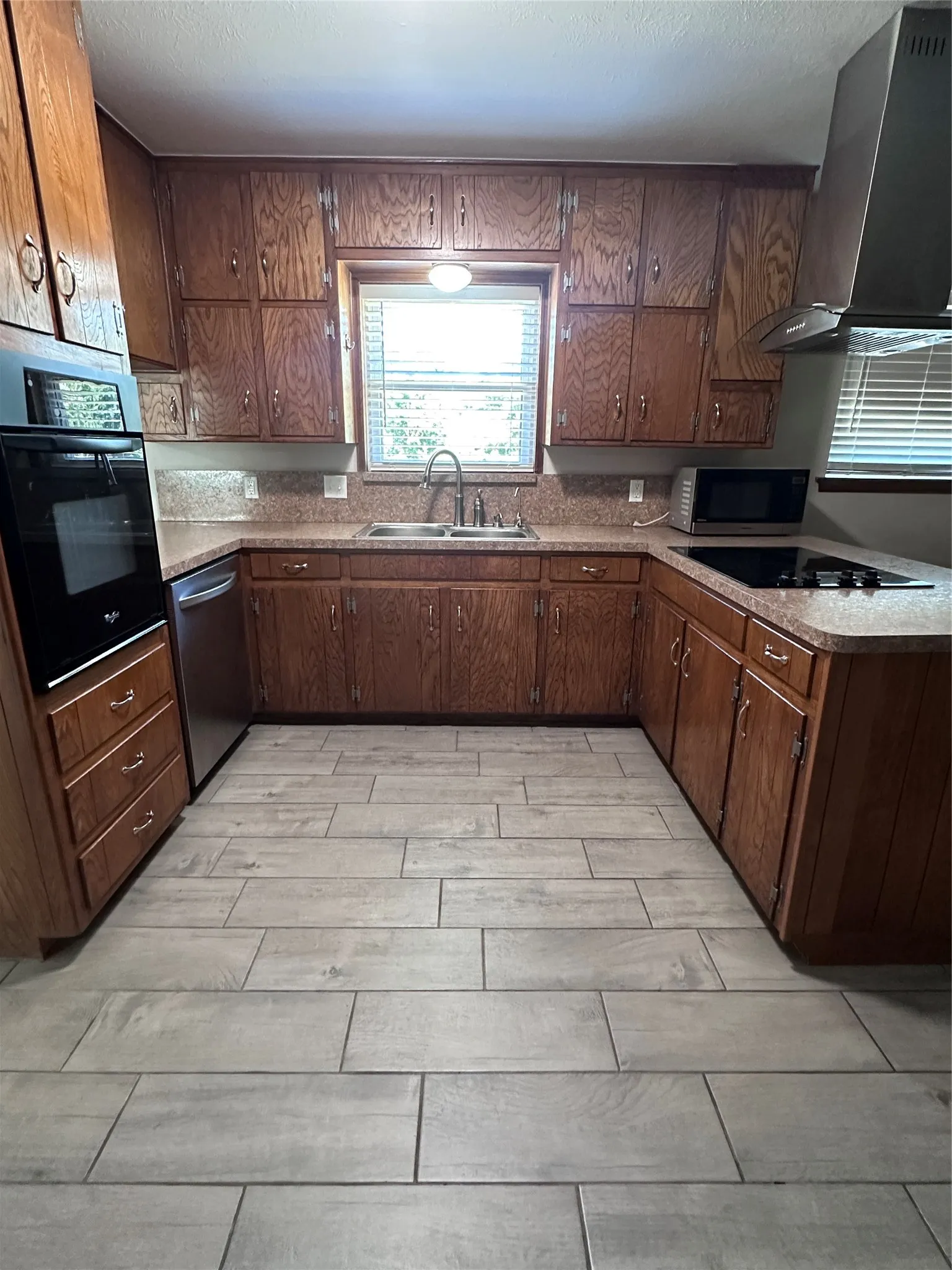 Wood-finish cabinetry, built-in oven, stainless steel dishwasher, kitchen sink with a gooseneck faucet, and light-toned tile flooring