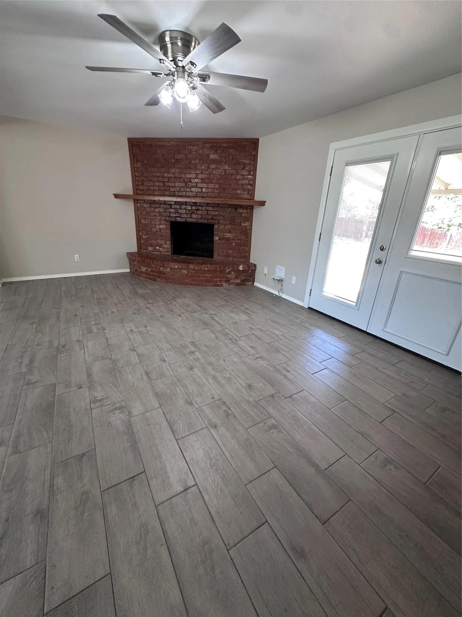Wood-finish flooring throughout, brick fireplace with a wood mantel, ceiling fan with integrated lighting, and French doors