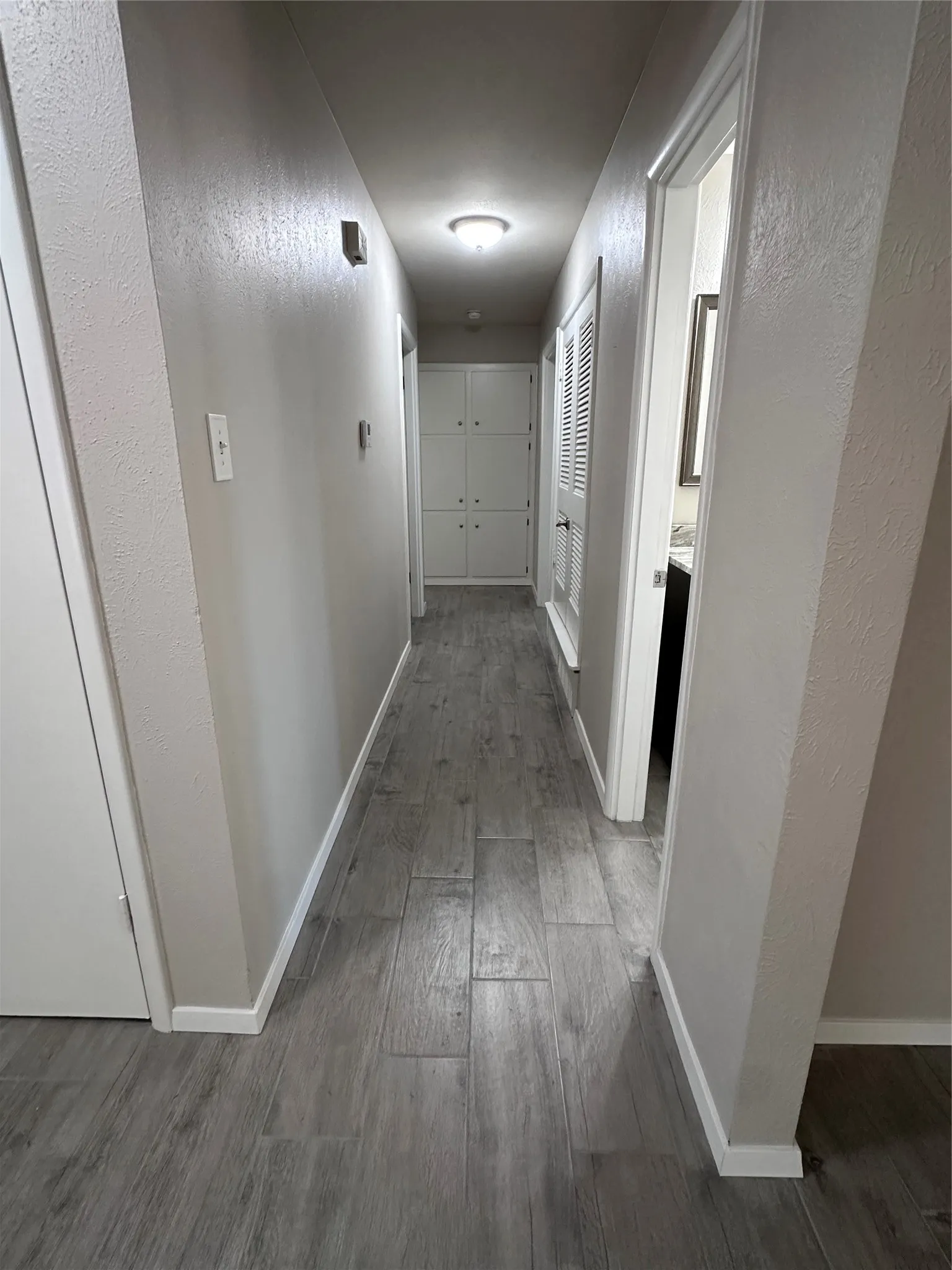 Recessed lighting illuminates the hallway, featuring wood-look plank tile flooring, built-in cabinetry, and a louvered door