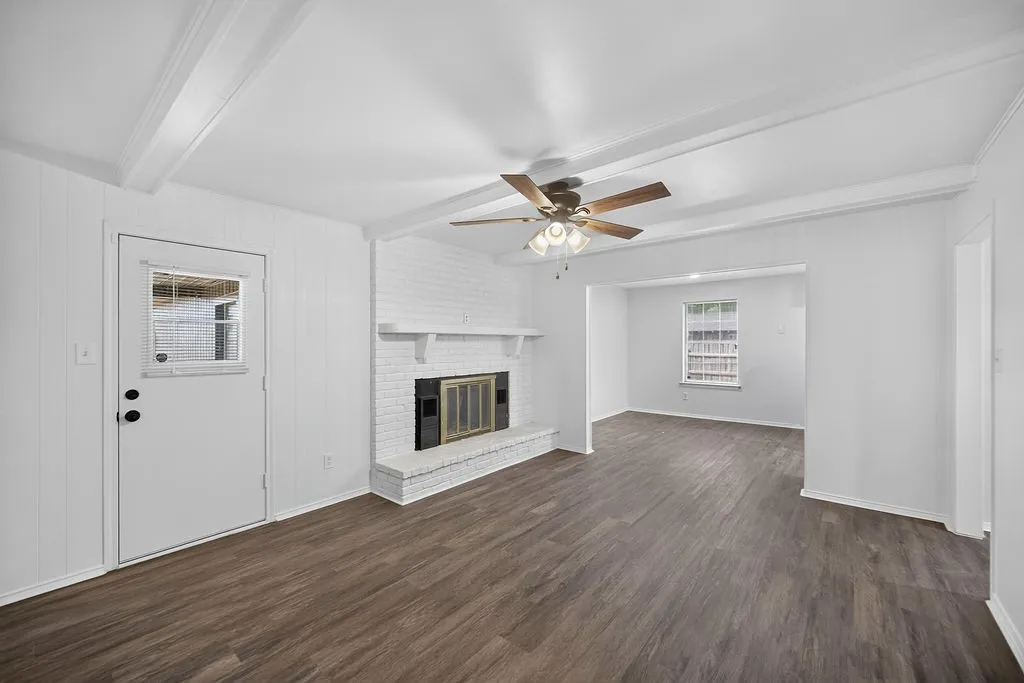 Wood-finish flooring throughout, white painted brick fireplace with hearth, exposed ceiling beams, and a ceiling fan with light fixture