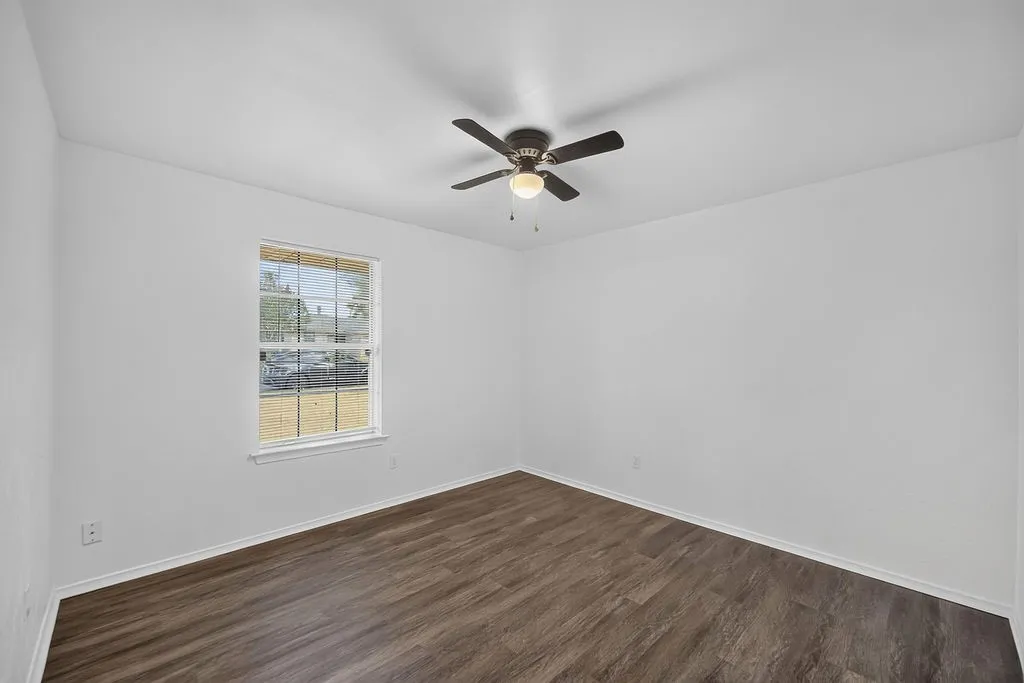 Wood-finish flooring, white walls, ceiling fan with integrated lighting, and a single window with white trim