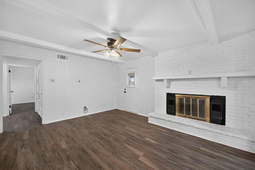 Wood-finish flooring, white painted brick fireplace with a brass firebox, white wood paneling, ceiling fan with integrated lighting, and white exposed ceiling beams