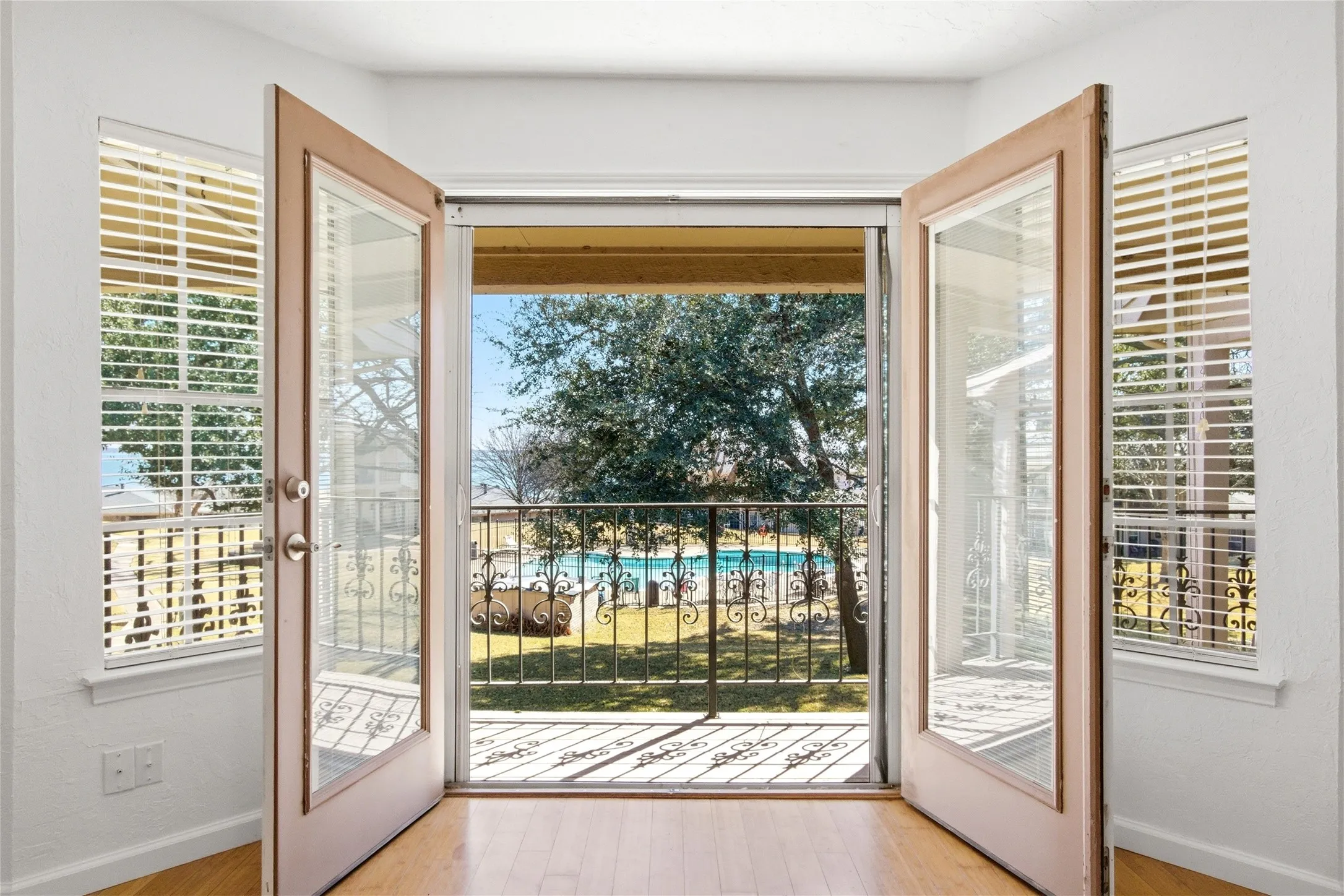 French doors opening to a balcony with decorative iron railing, overlooking a pool and mature trees