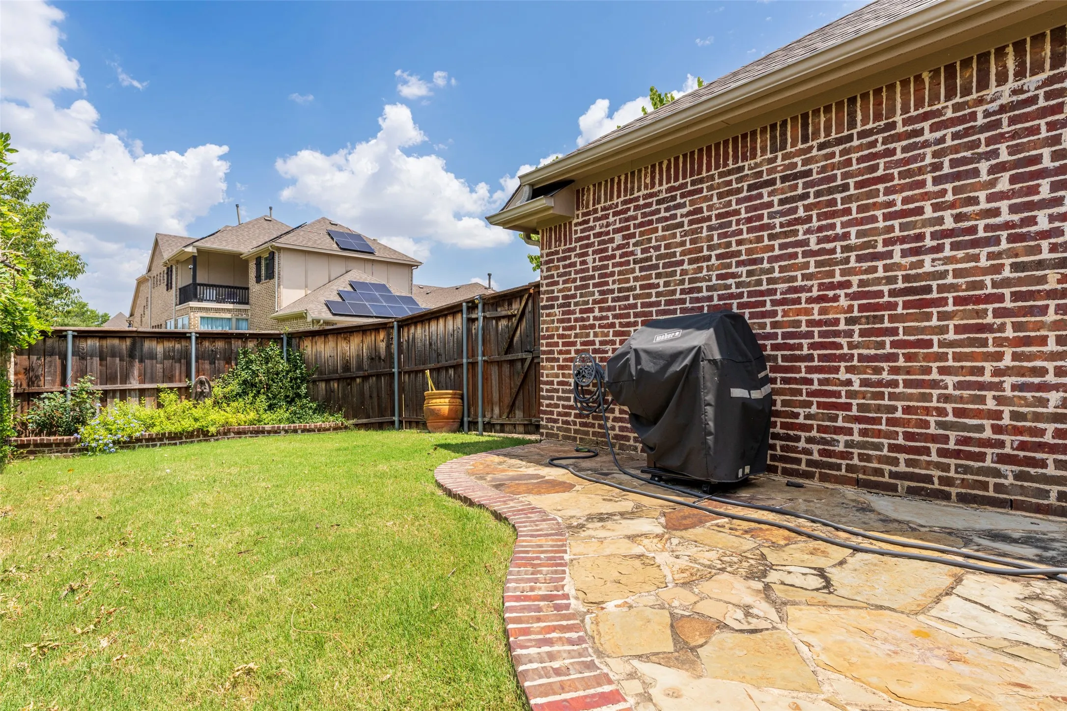Low-maintenance backyard with mature landscaping, grassy space, and an open flagstone patio accented by brick edging. Capped wood privacy fencing provides a defined outdoor space for pets, gardening, and entertaining.